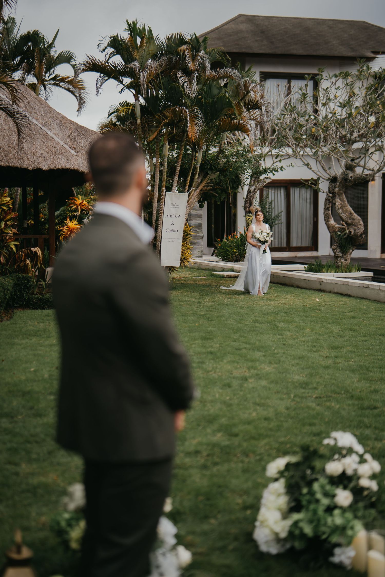 Groom watches tattooed bride walk across lush garden lawn at tropical Bali wedding venue.