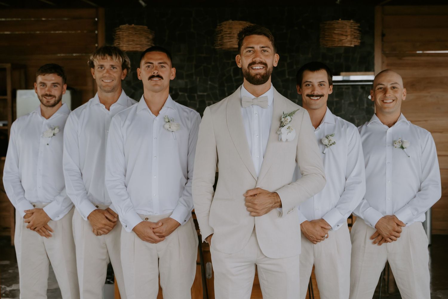 Groom in beige suit stands with five groomsmen all wearing white shirts and mustaches outdoors.