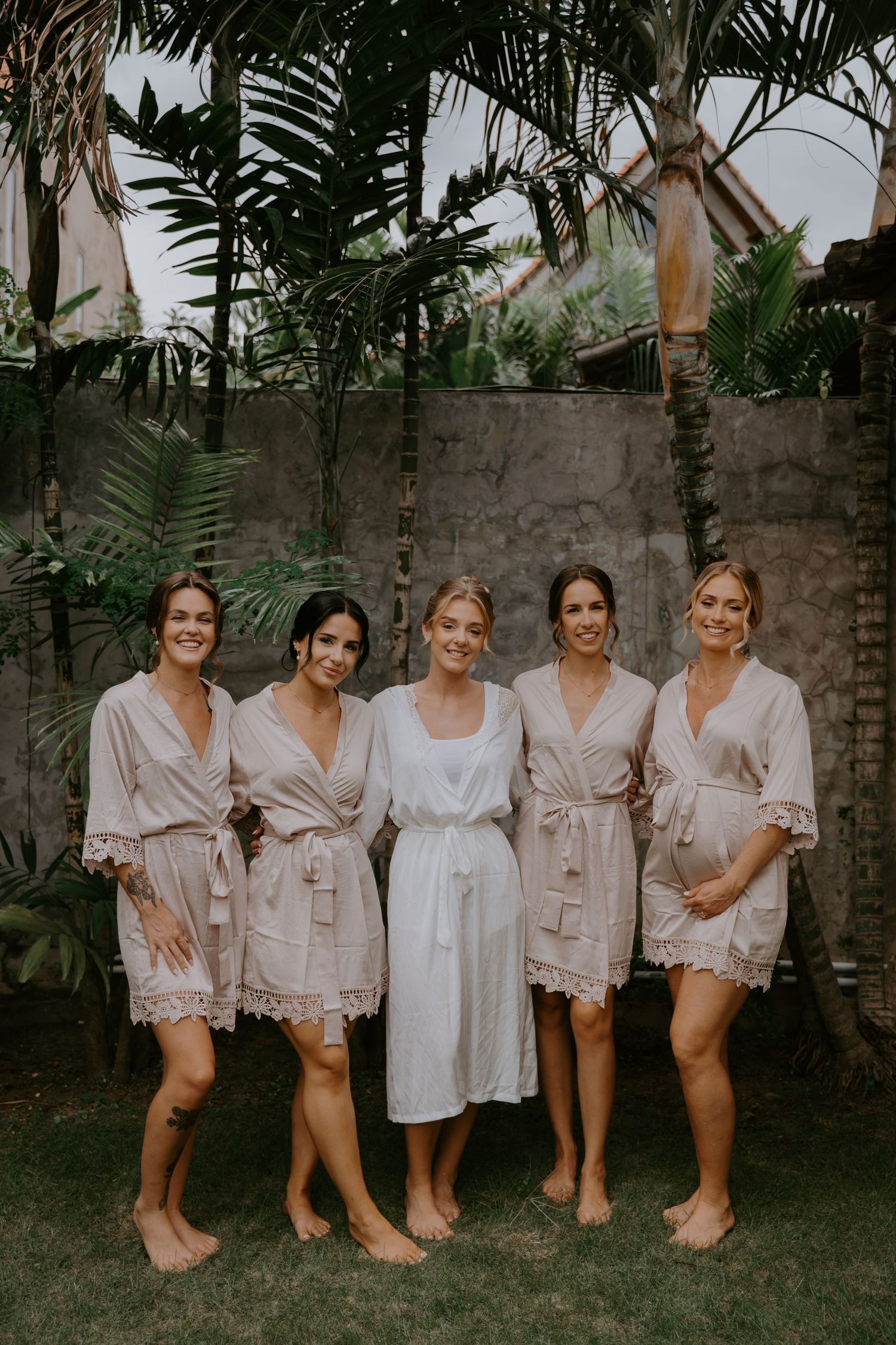 Bride in white robe poses with four bridesmaids in blush robes in tropical garden setting.