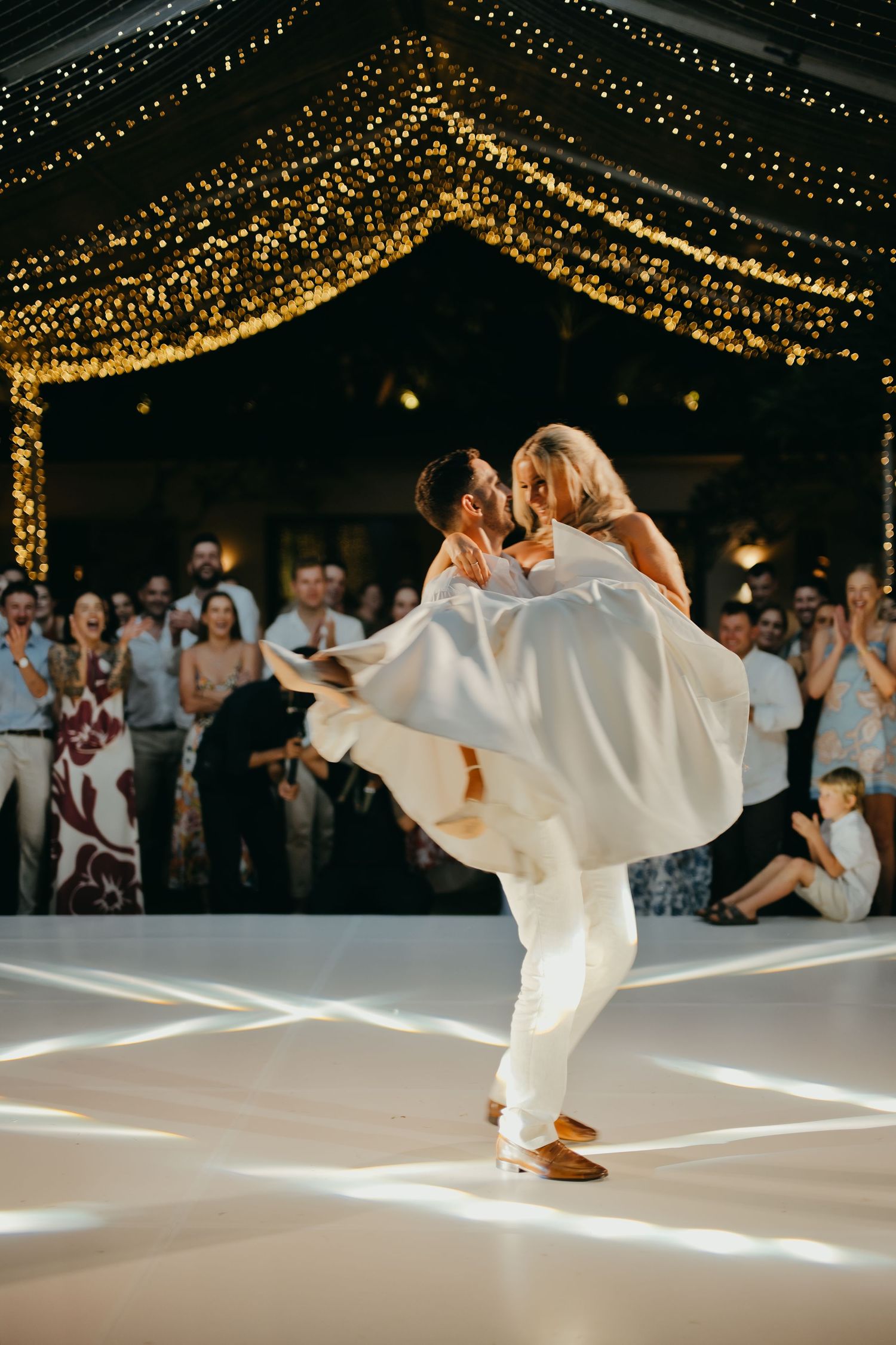 Groom lifts bride in a sweeping romantic moment on a white dance floor at their fairy-lit wedding reception.