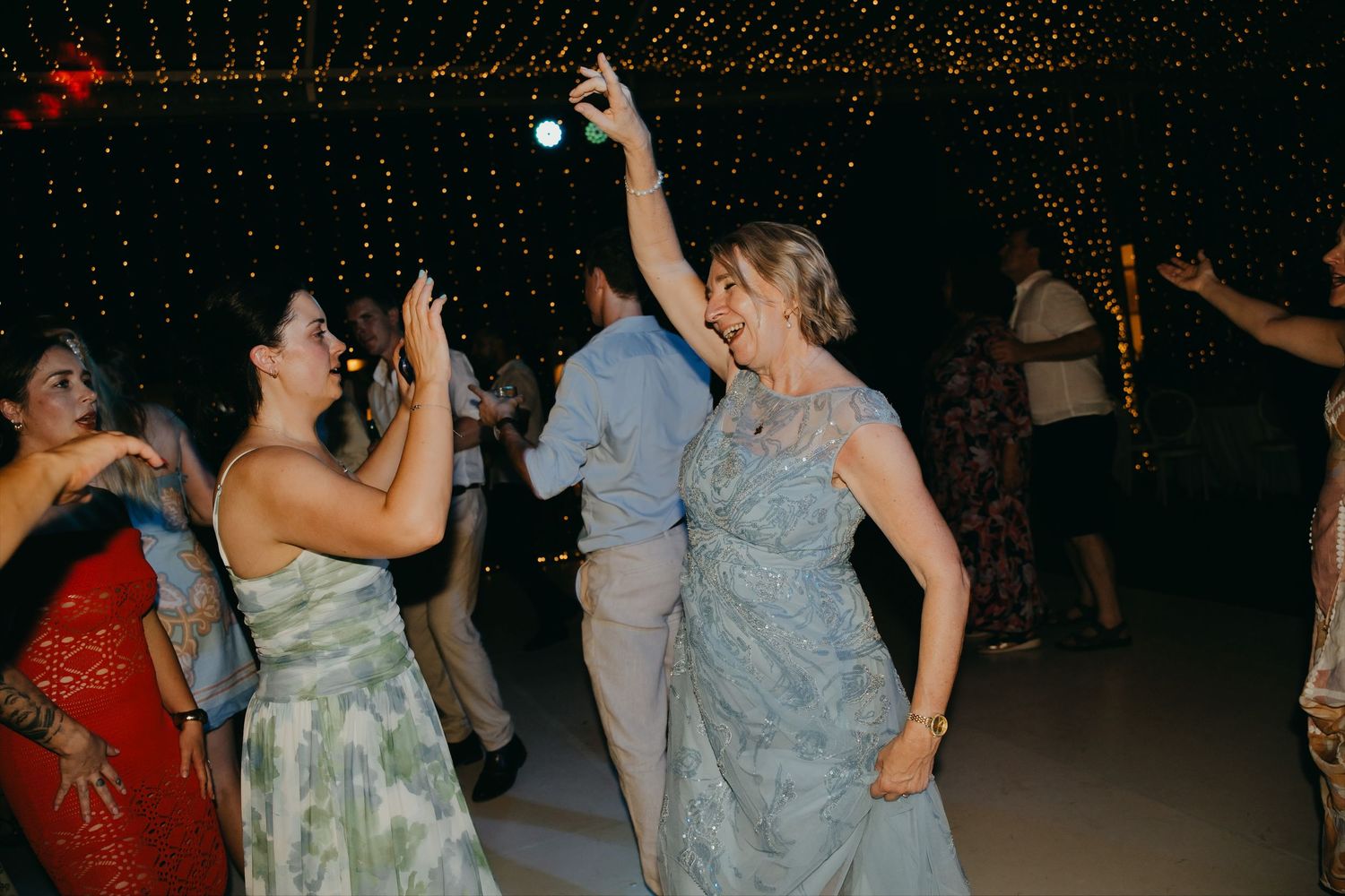Two women laughing and dancing together under twinkling fairy lights at a wedding reception.