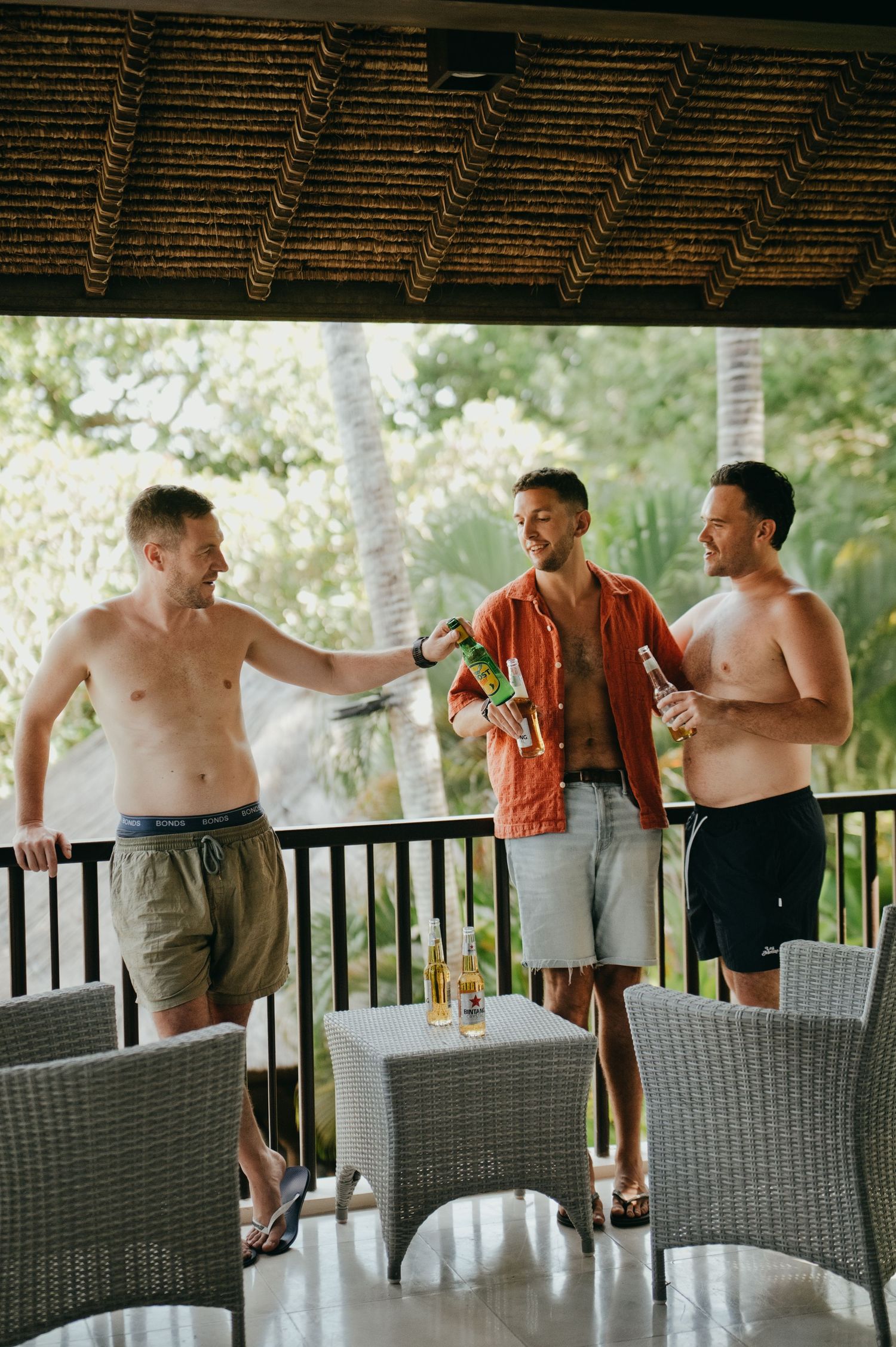 Three men socializing on tropical villa terrace with beer bottles under thatched roof.