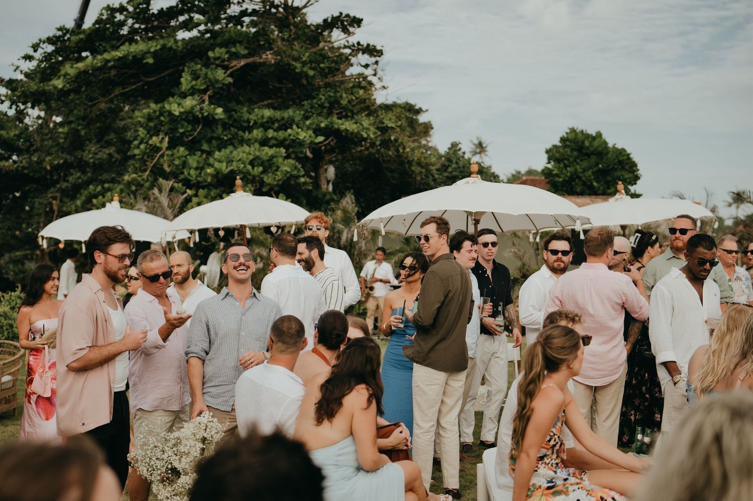 Wedding guests mingling outdoors under white parasols at a tropical garden cocktail reception.