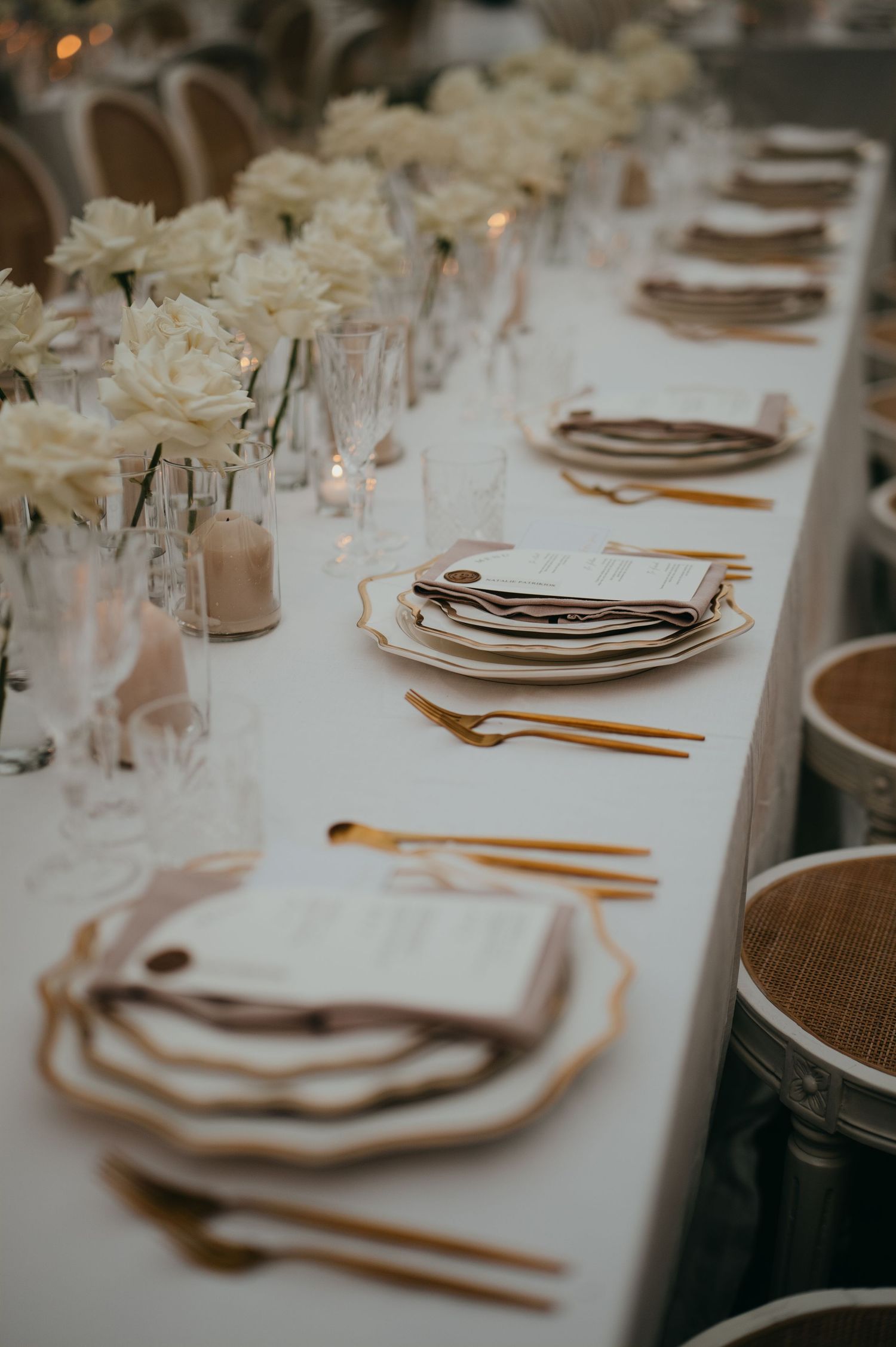 Elegant wedding reception table with gold cutlery, white floral centerpieces, and layered place settings on white linen.
