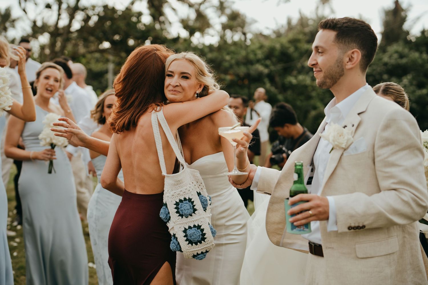 Groom in ivory suit hugging a guest while others celebrate at an outdoor tropical wedding reception.