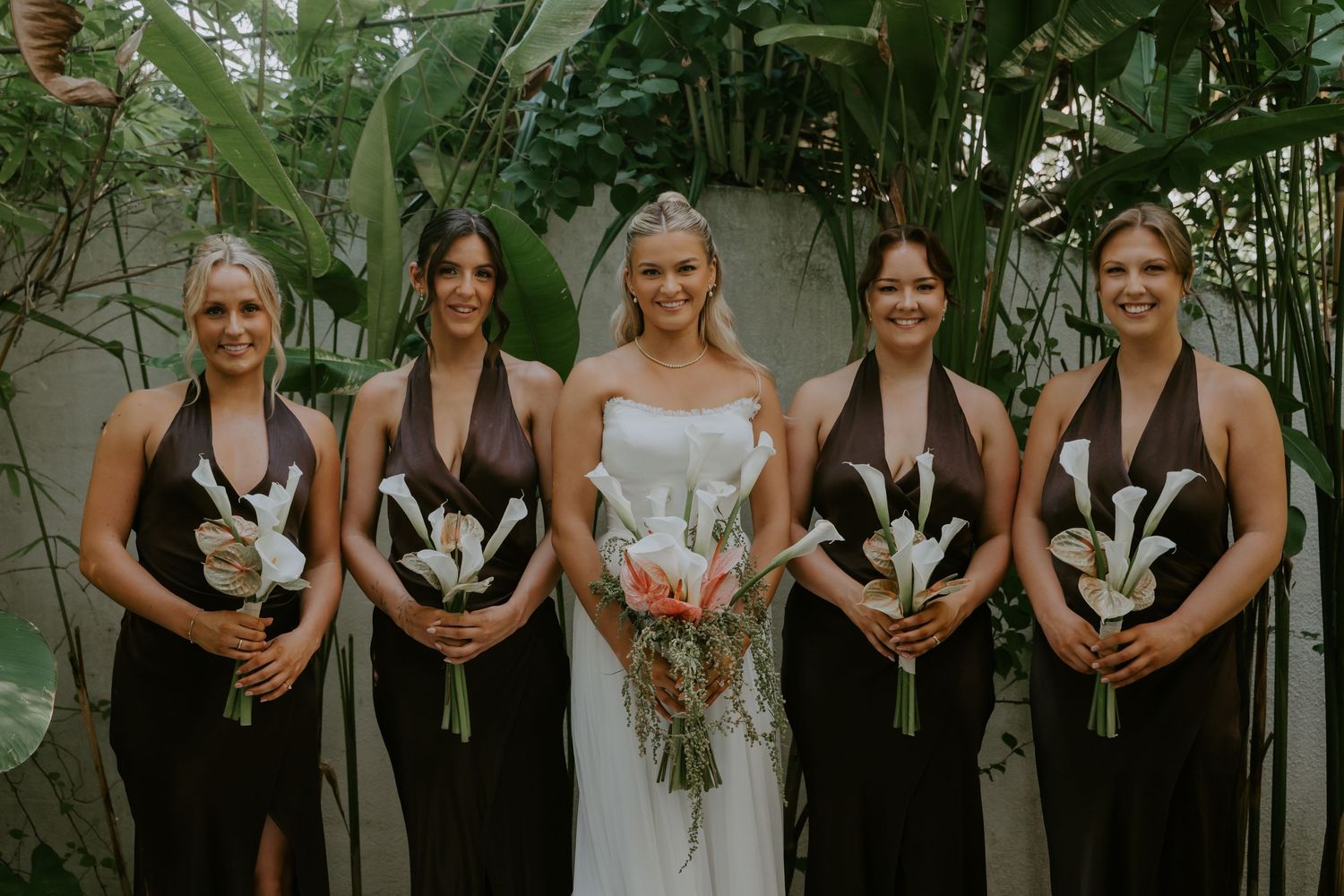 Bride holding tropical bouquet poses with four bridesmaids in dark brown halter dresses among lush greenery.