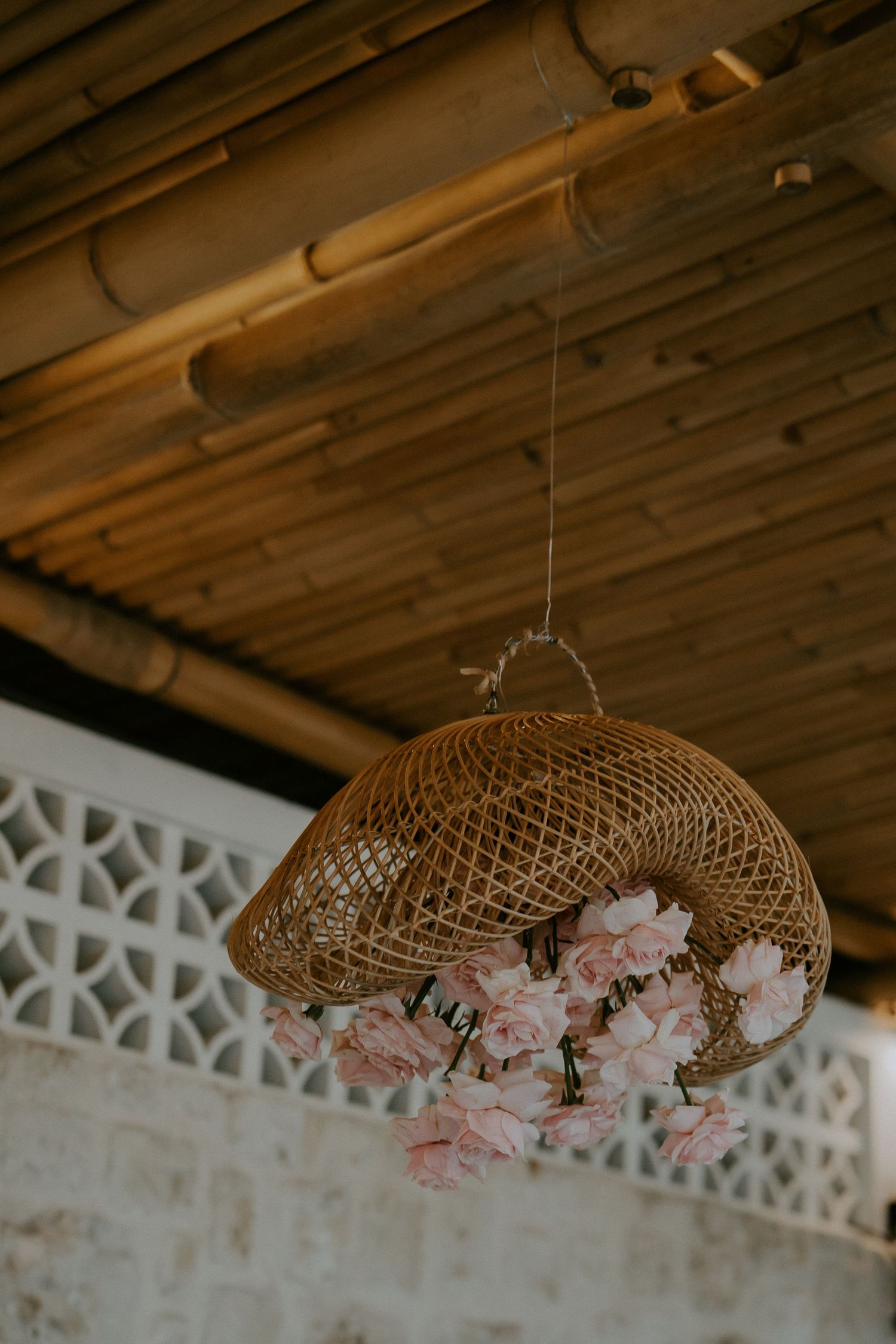 Decorative wicker pendant light adorned with pink roses hanging from rustic bamboo ceiling at wedding venue.