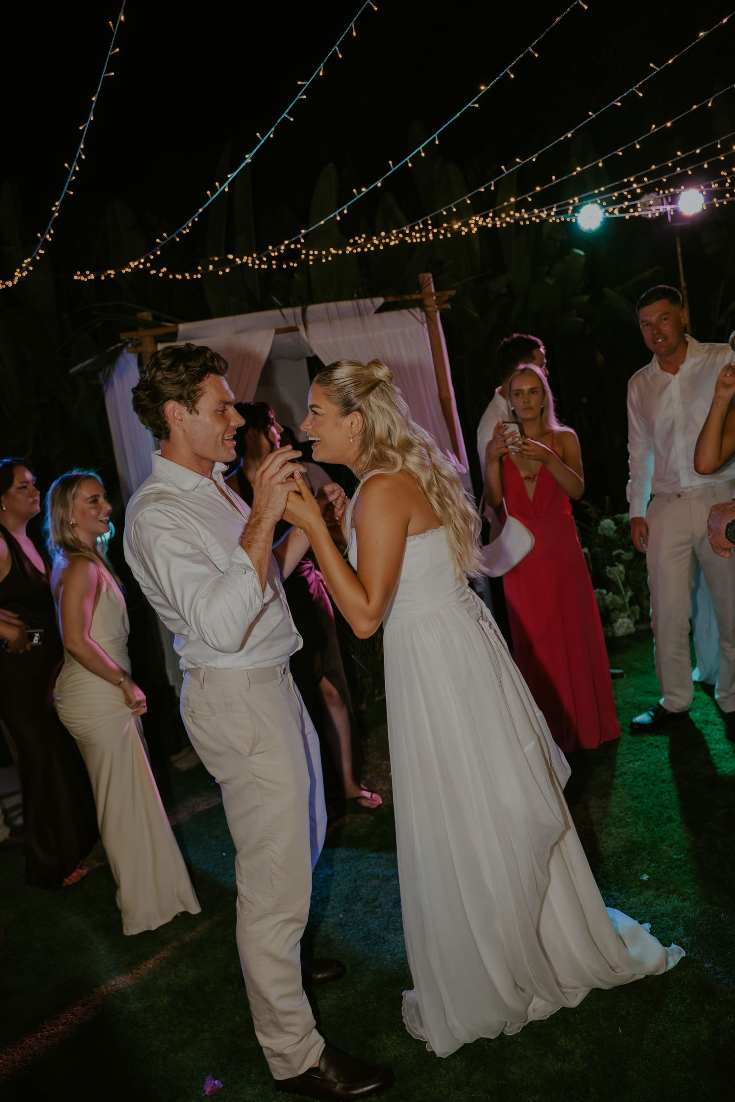 Bride and groom sharing their first dance outdoors under fairy lights with guests watching.