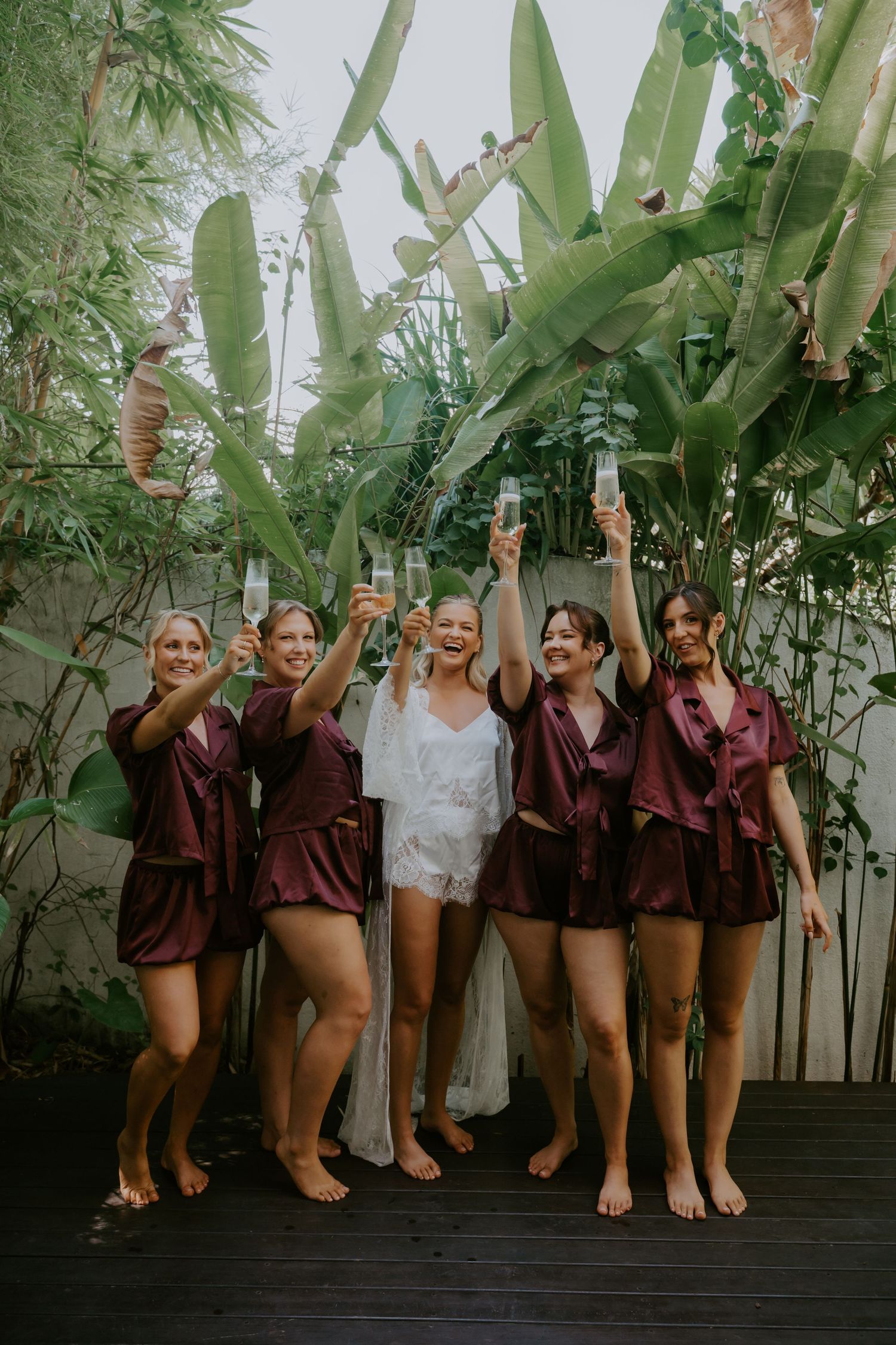 Bride in white lace robe toasting champagne with bridesmaids in burgundy satin sets surrounded by tropical greenery.