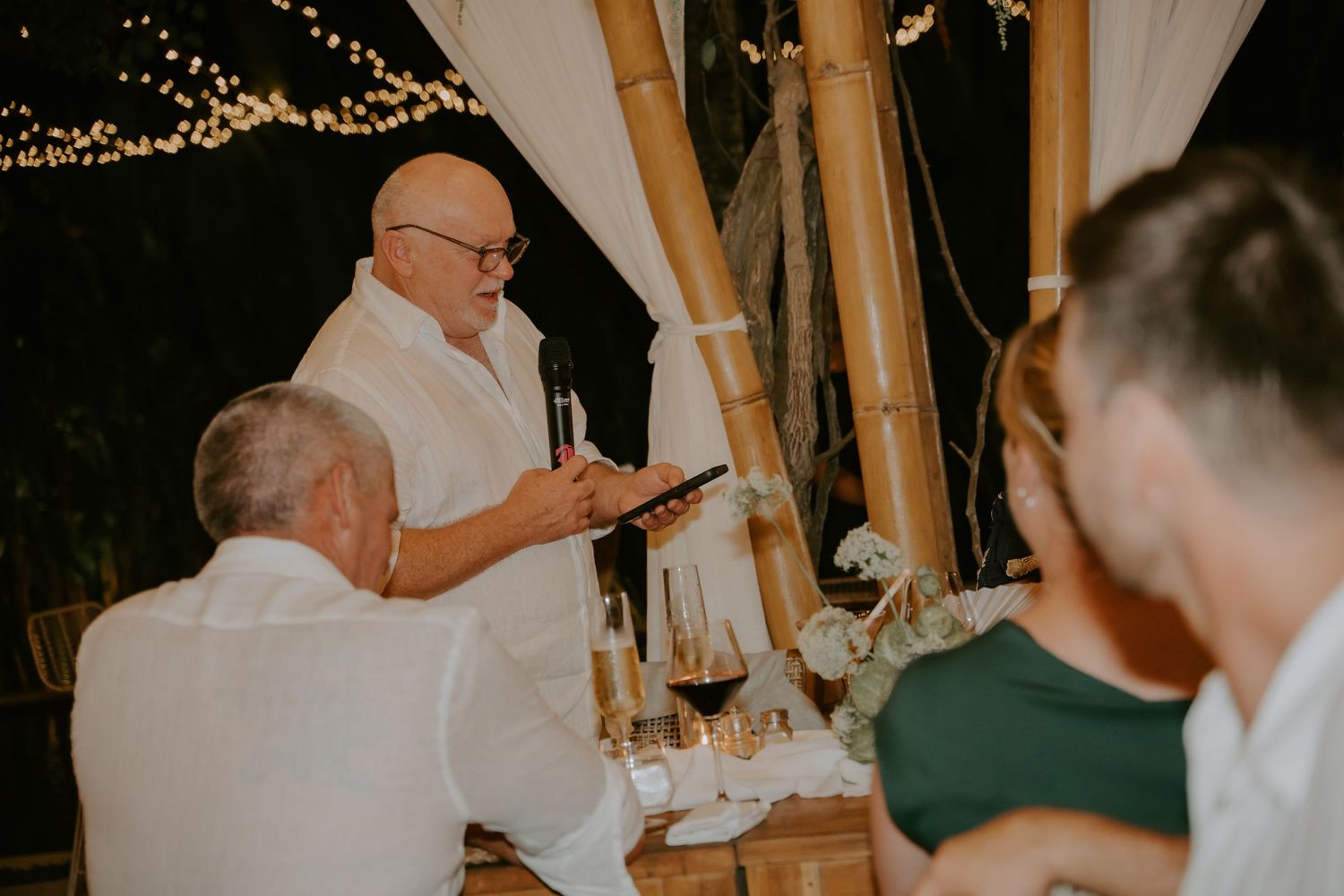 Elderly man giving a heartfelt wedding speech at an outdoor bamboo-decorated reception venue.
