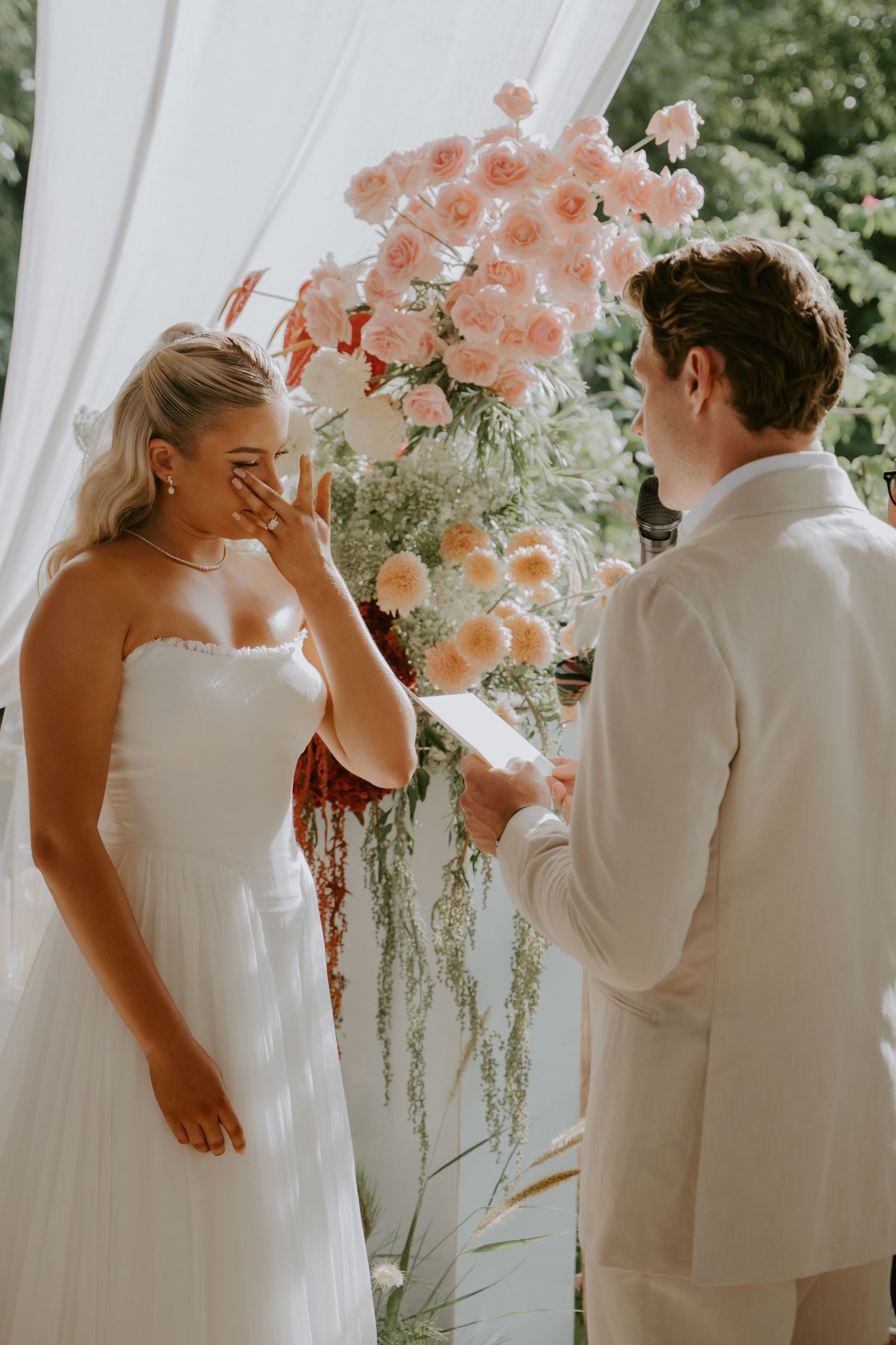 Emotional bride wiping tears as groom reads heartfelt vows at a floral-adorned outdoor wedding ceremony.