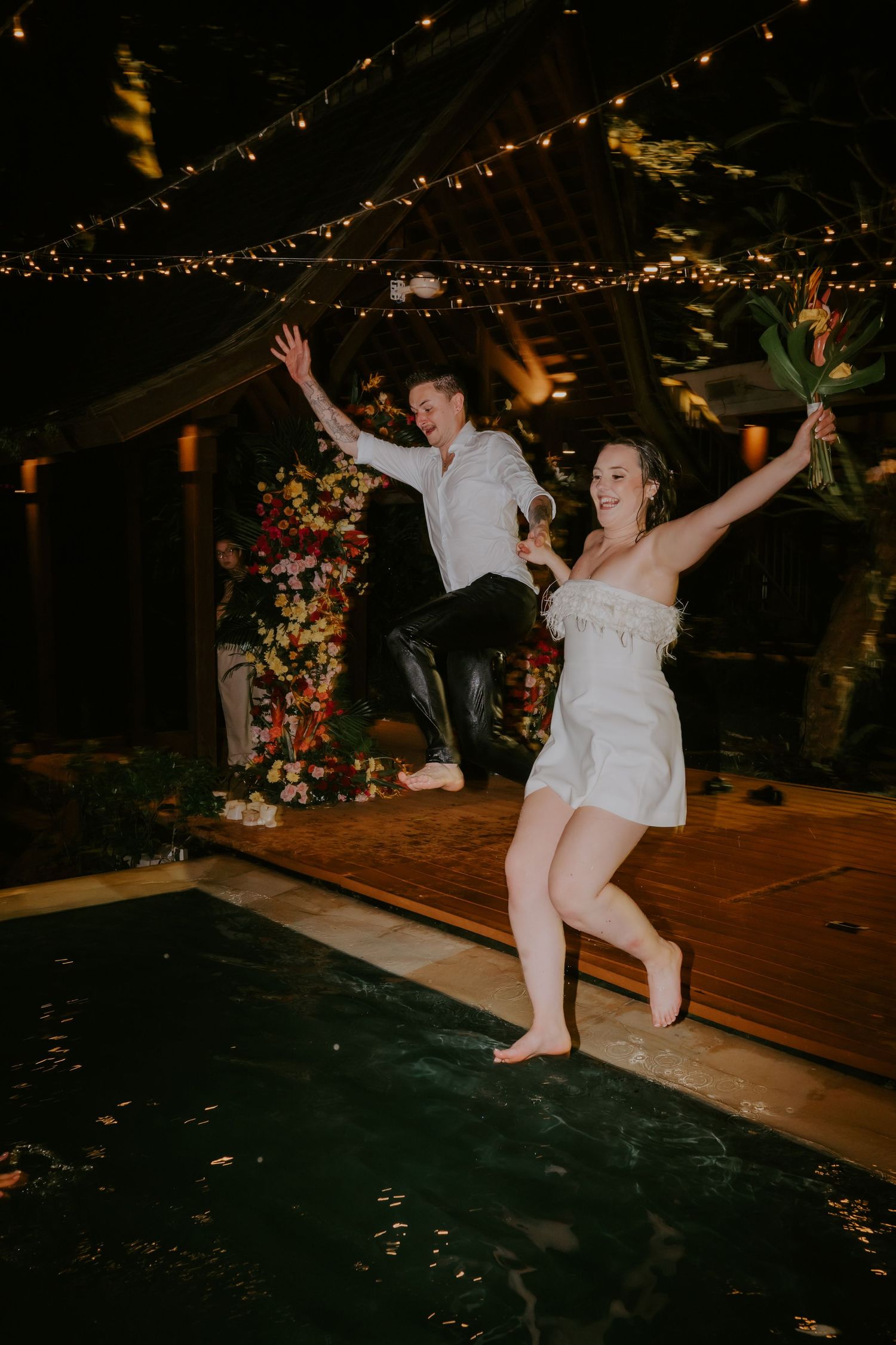 Joyful bride and groom jumping into pool at night during wedding celebration with floral backdrop.