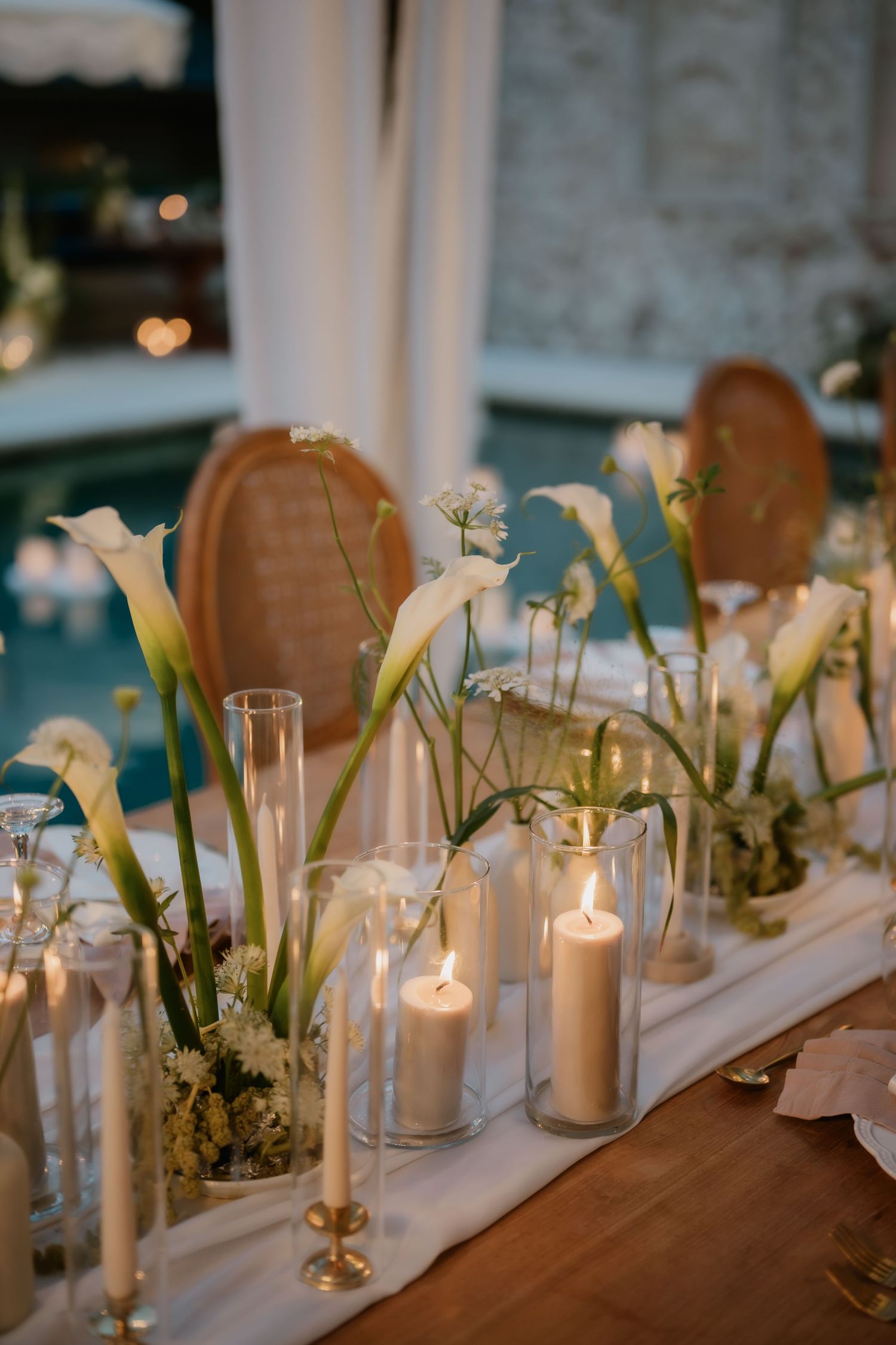 Elegant wedding reception table with white calla lilies, pillar candles in glass vases, and gold candlesticks on white runner.