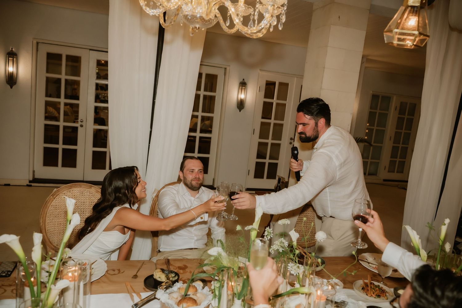 Groom toasts bride at intimate wedding reception dinner table decorated with white calla lilies.