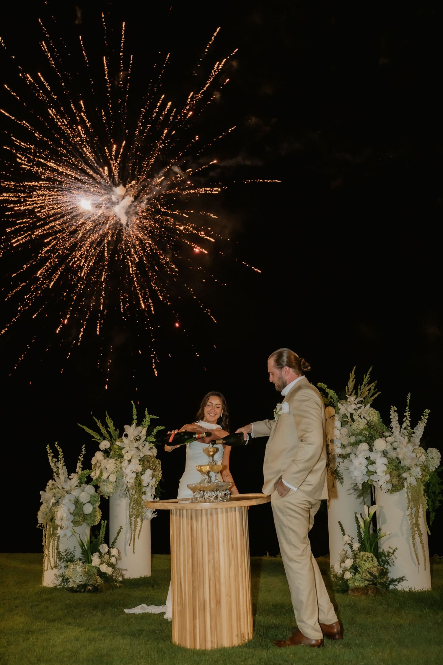 Couple cutting wedding cake with fireworks display behind white floral arrangements at night.