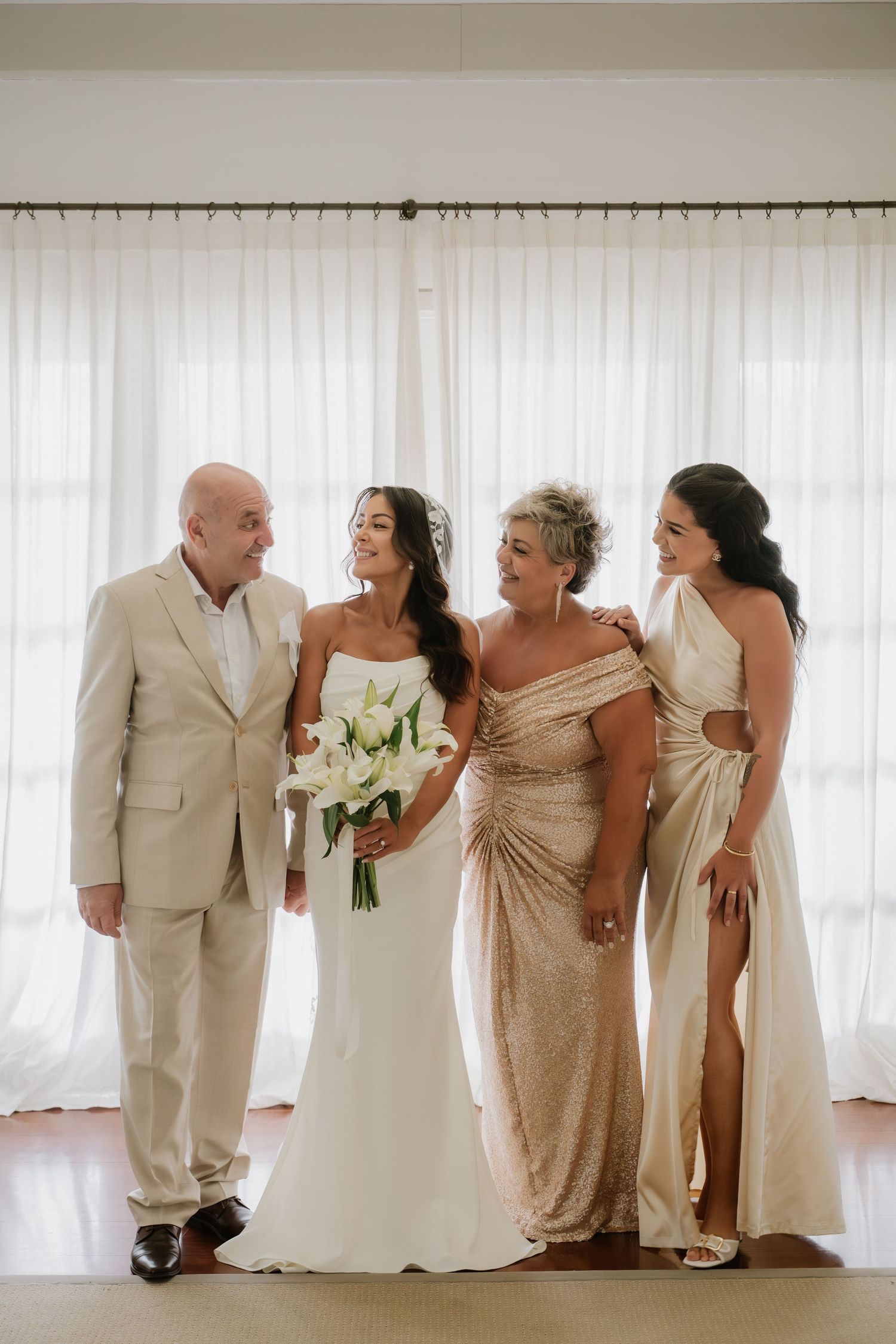 Bride in strapless white gown poses with family in neutral attire holding white lily bouquet indoors.