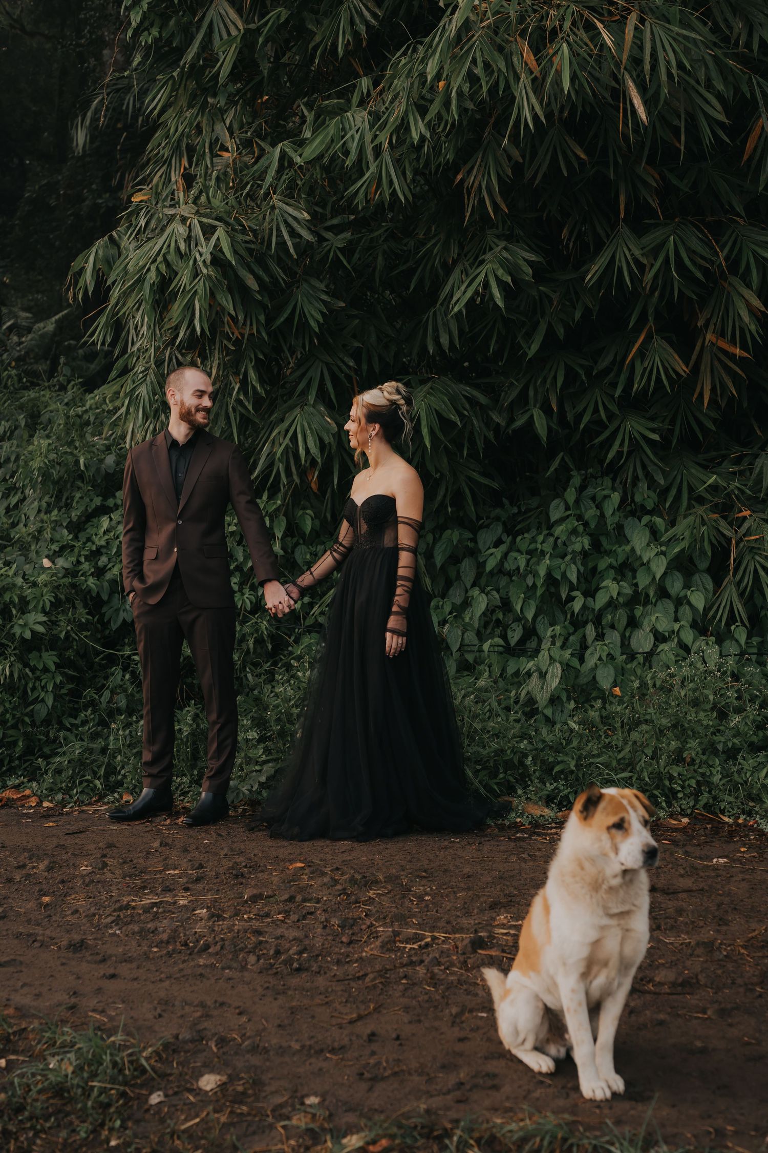 A couple poses with their dog in a lush garden setting with bamboo trees in the background.
