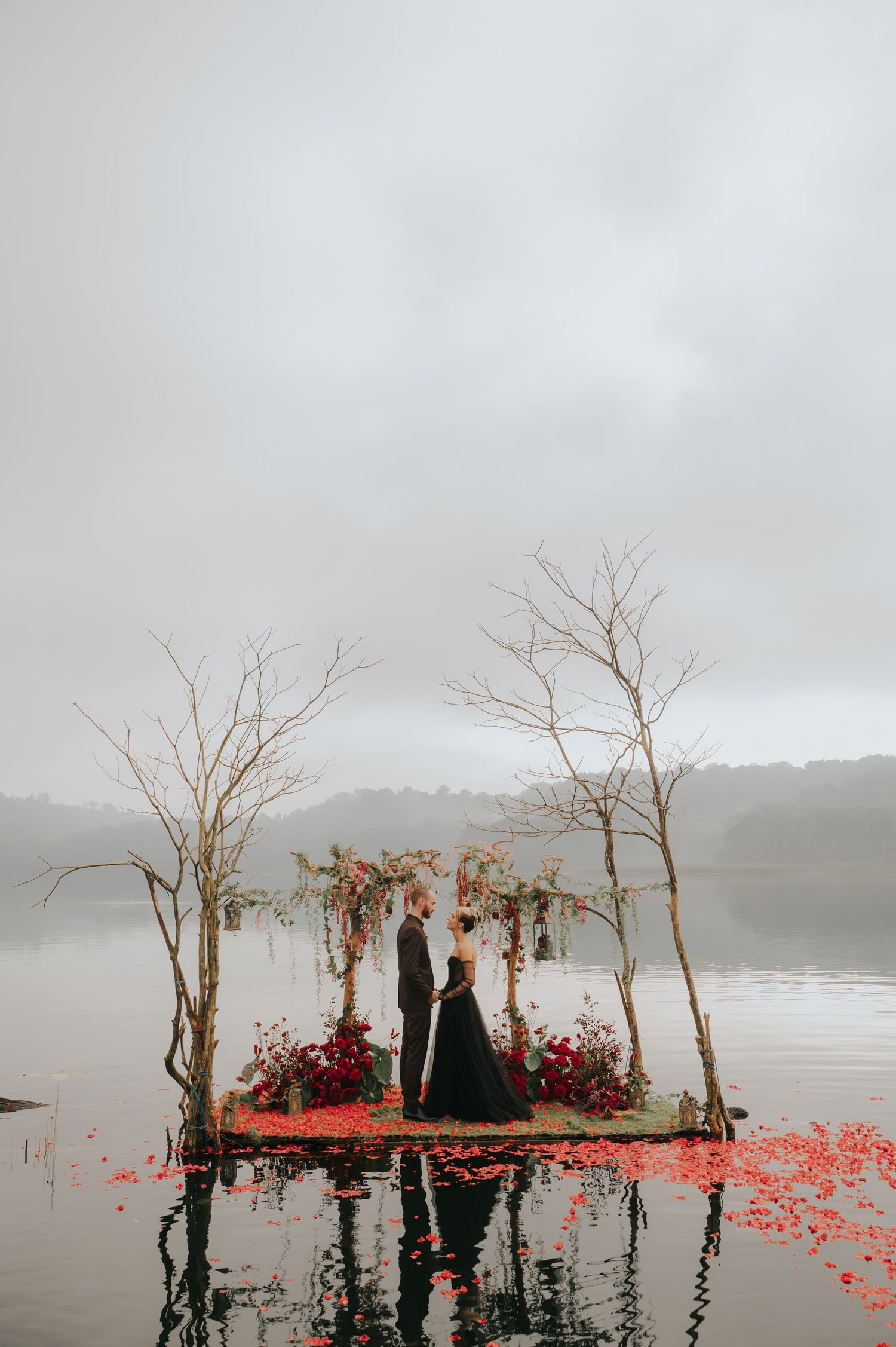 A floating wedding platform on misty water decorated with red flowers and bare trees.
