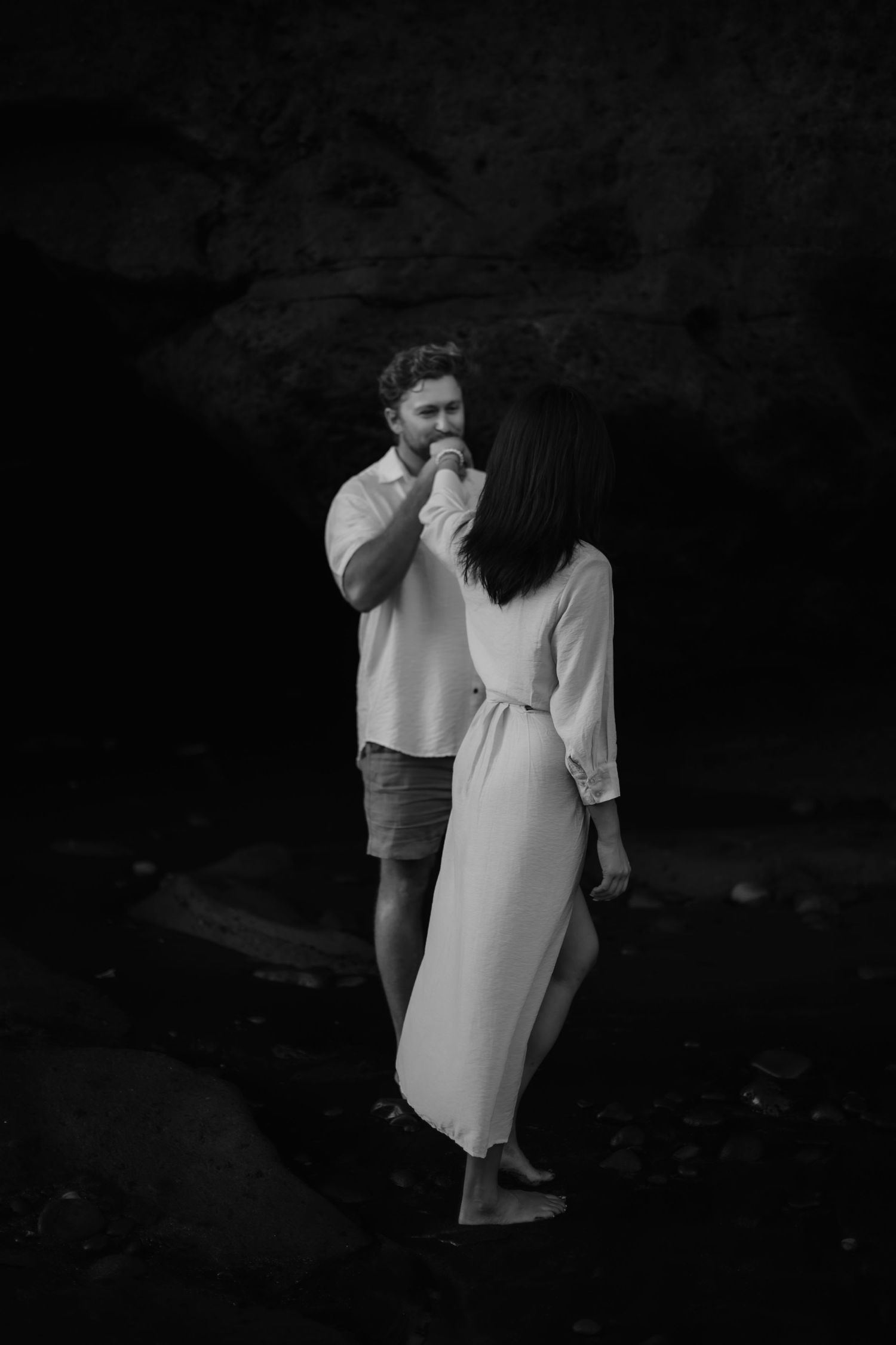 A black and white photo of a couple in white clothing standing together in a dark, moody setting.