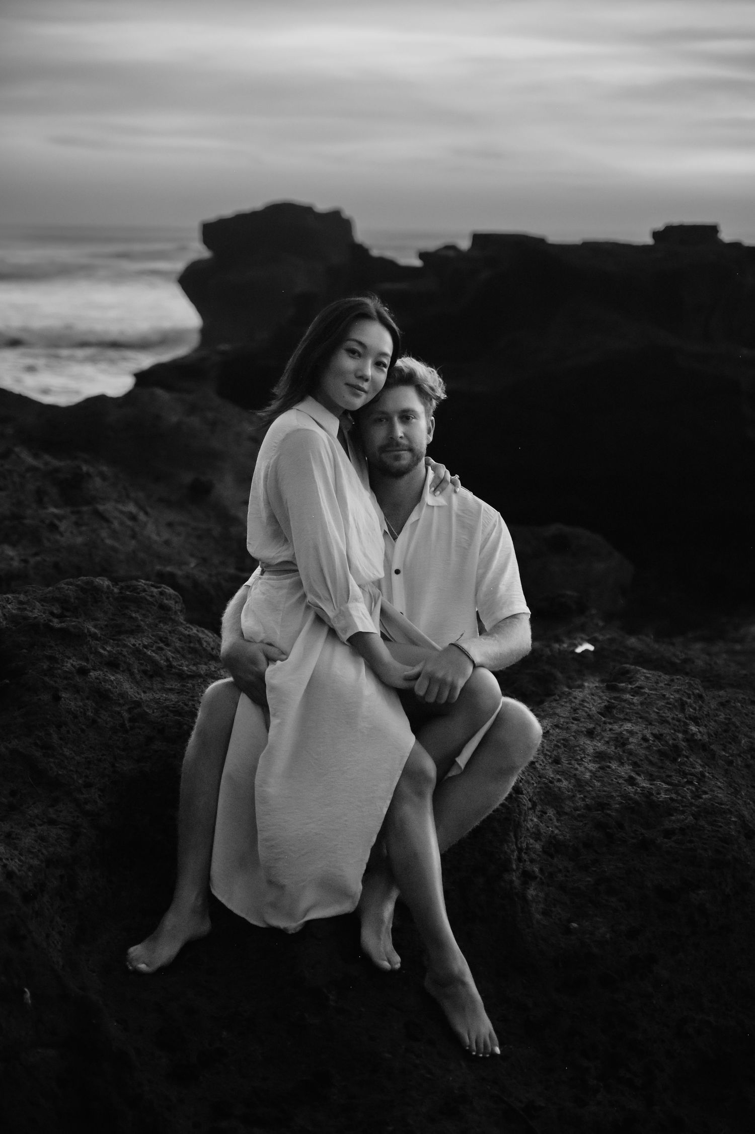 Black and white romantic beach photoshoot of couple sitting together on coastal rocks at sunset.