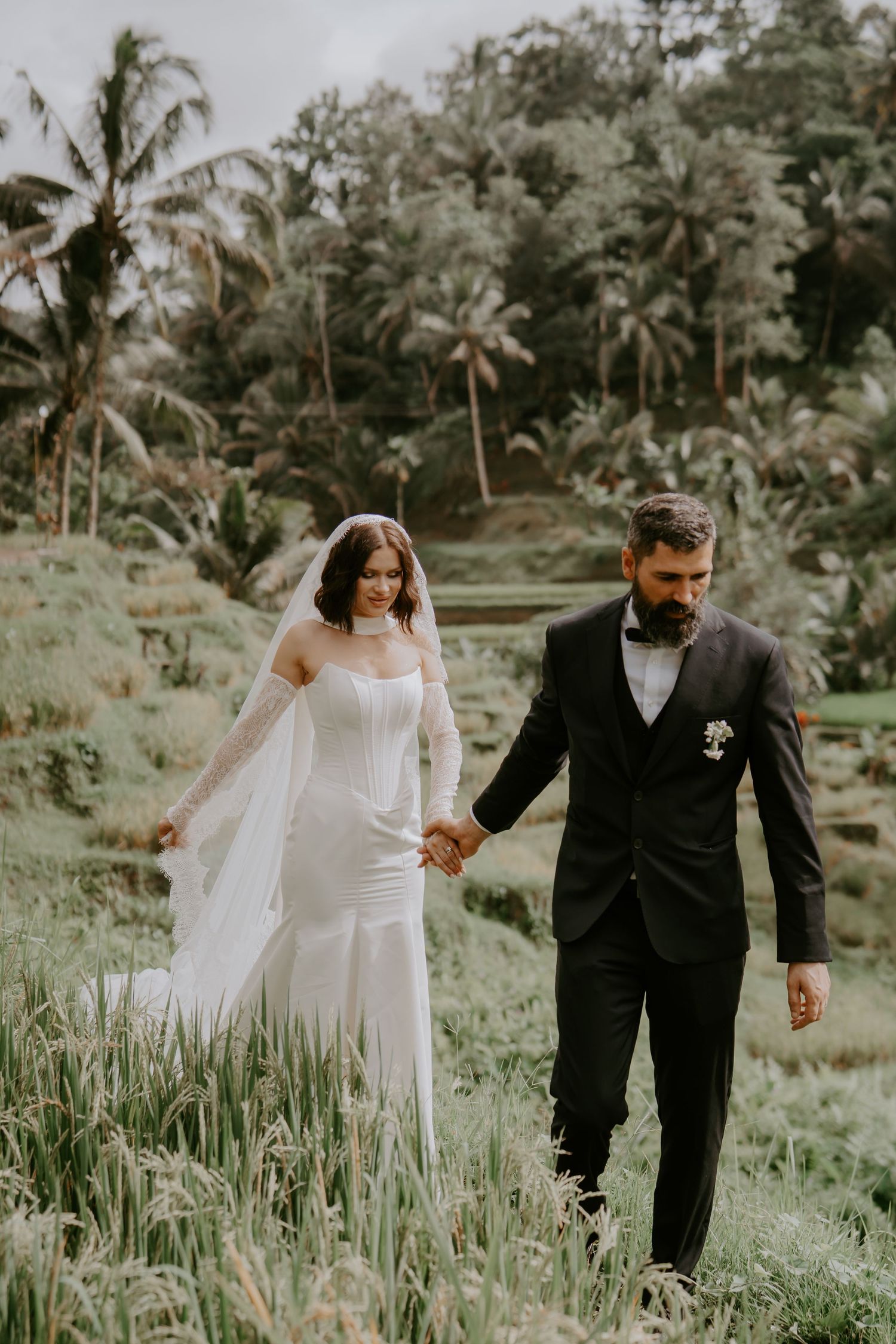 A couple in wedding attire walks hand in hand through a lush tropical garden setting with palm trees in the background.