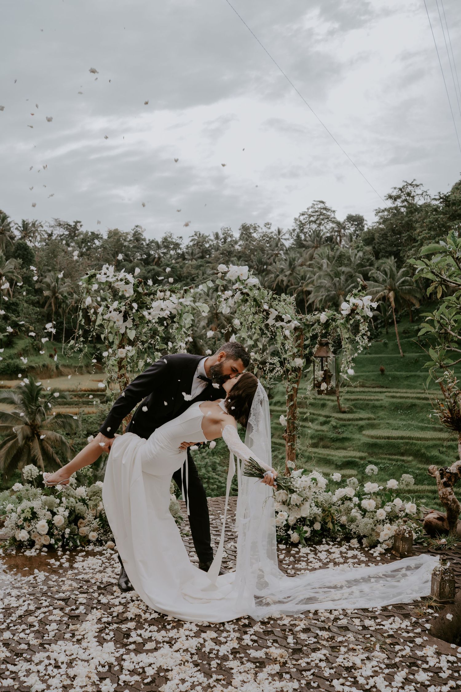 A romantic wedding photo captured in a lush tropical garden with terraced rice fields and floating flower petals in Bali.