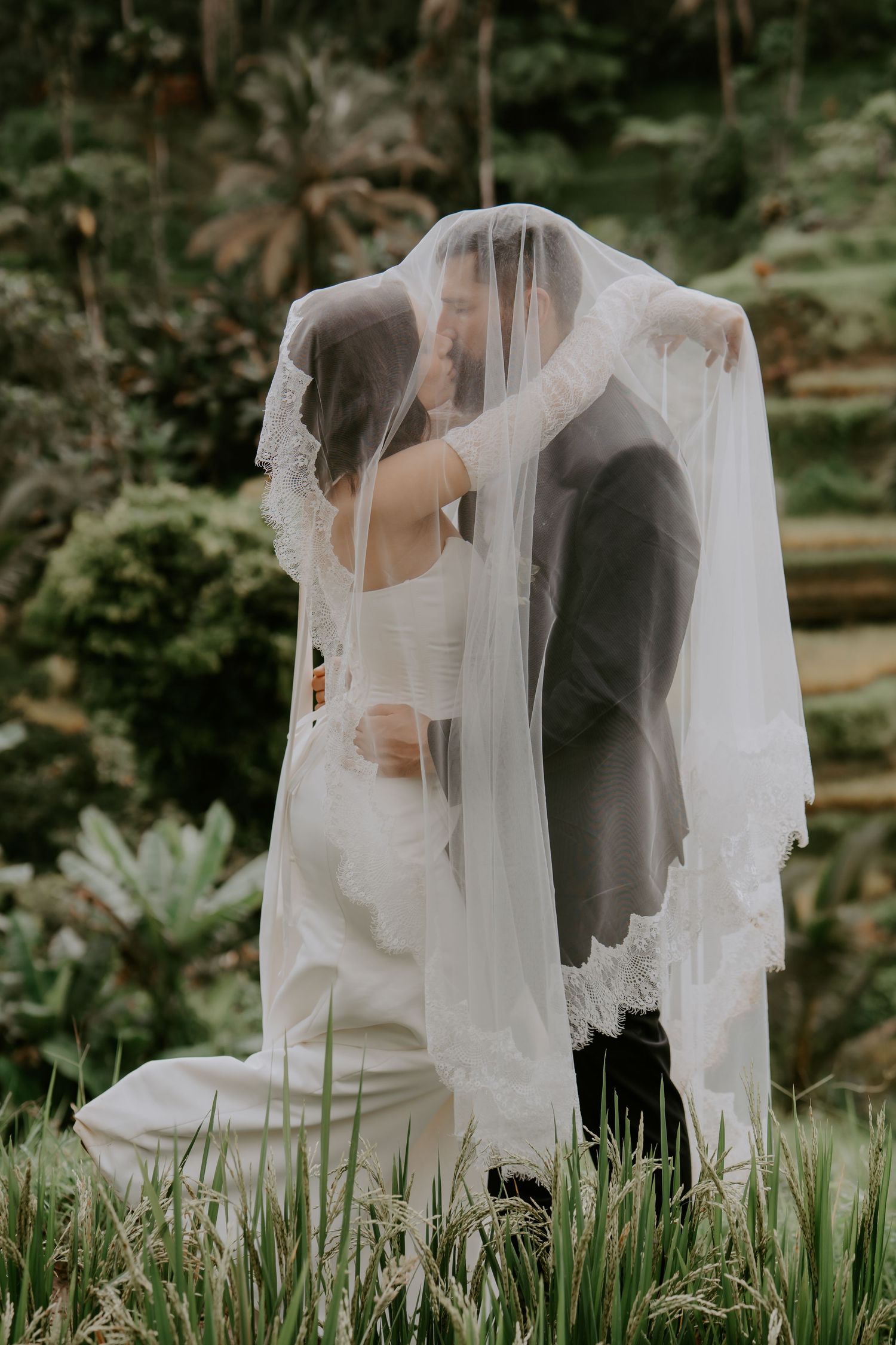 A romantic wedding couple sits together in a garden setting with flowing white veil creating an ethereal atmosphere.