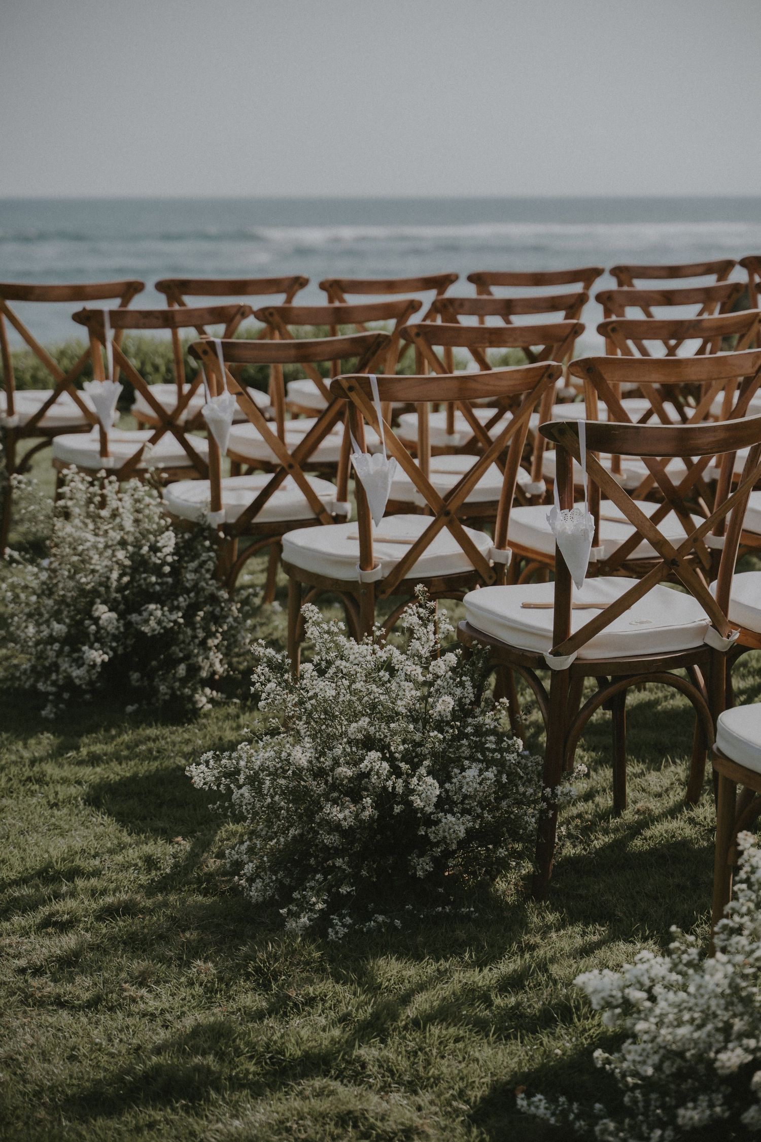 Rustic wooden cross-back chairs arranged for an oceanside wedding ceremony with delicate white flowers lining the aisle.
