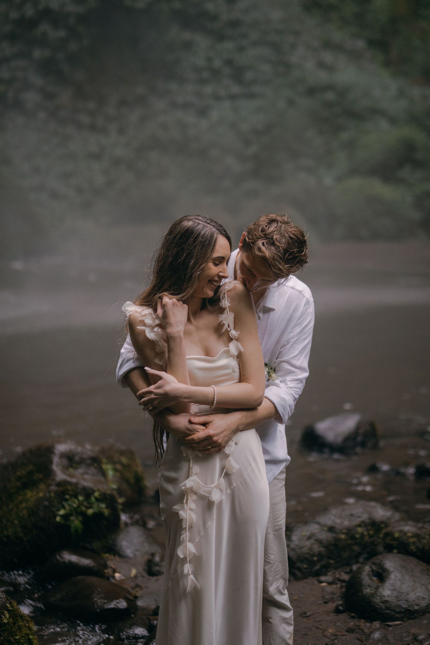 A couple in wedding attire shares an intimate embrace in a misty outdoor setting with rocky terrain.