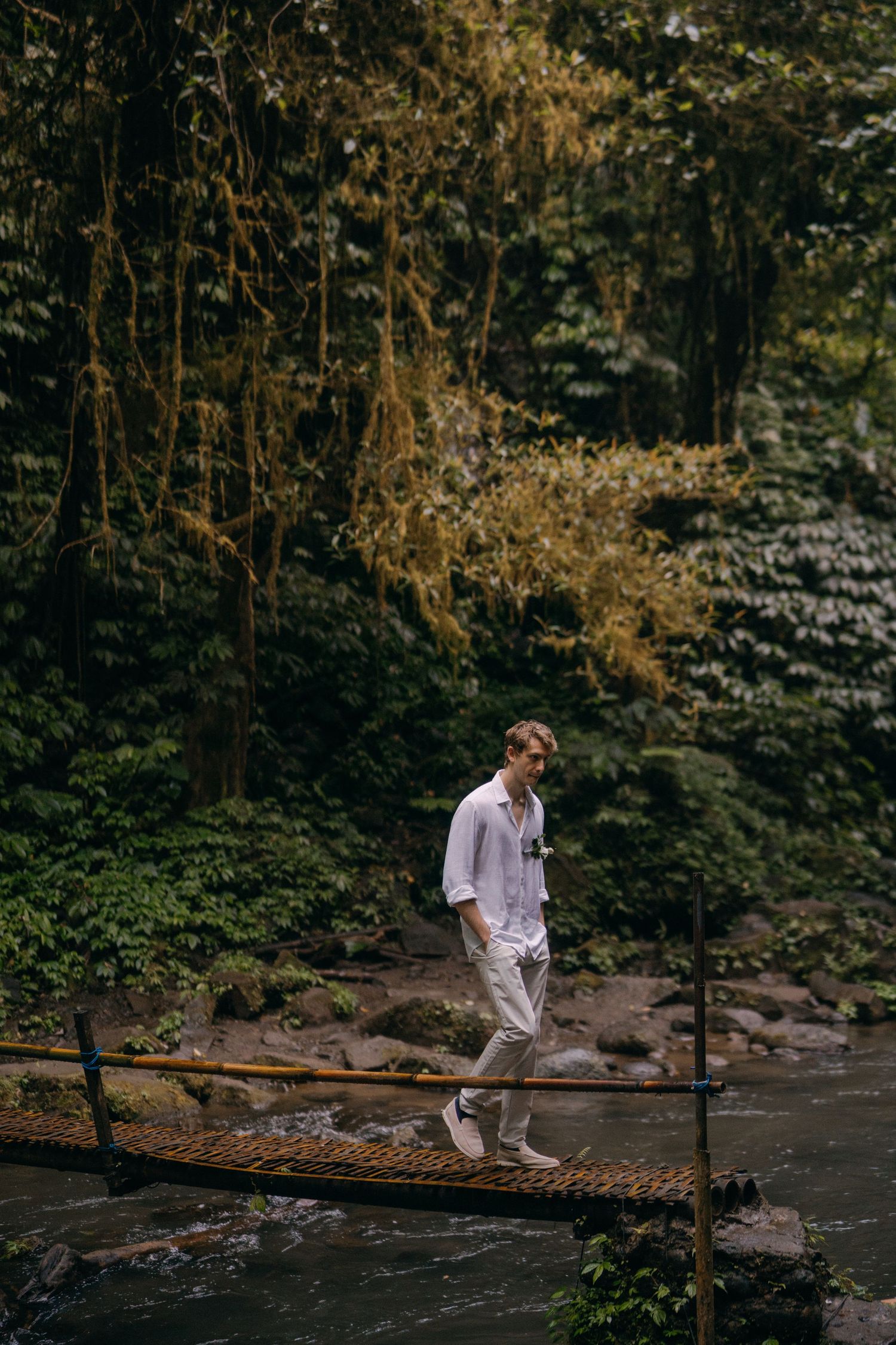 A person in white clothing walks across a wooden suspension bridge over a stream in a lush forest setting.