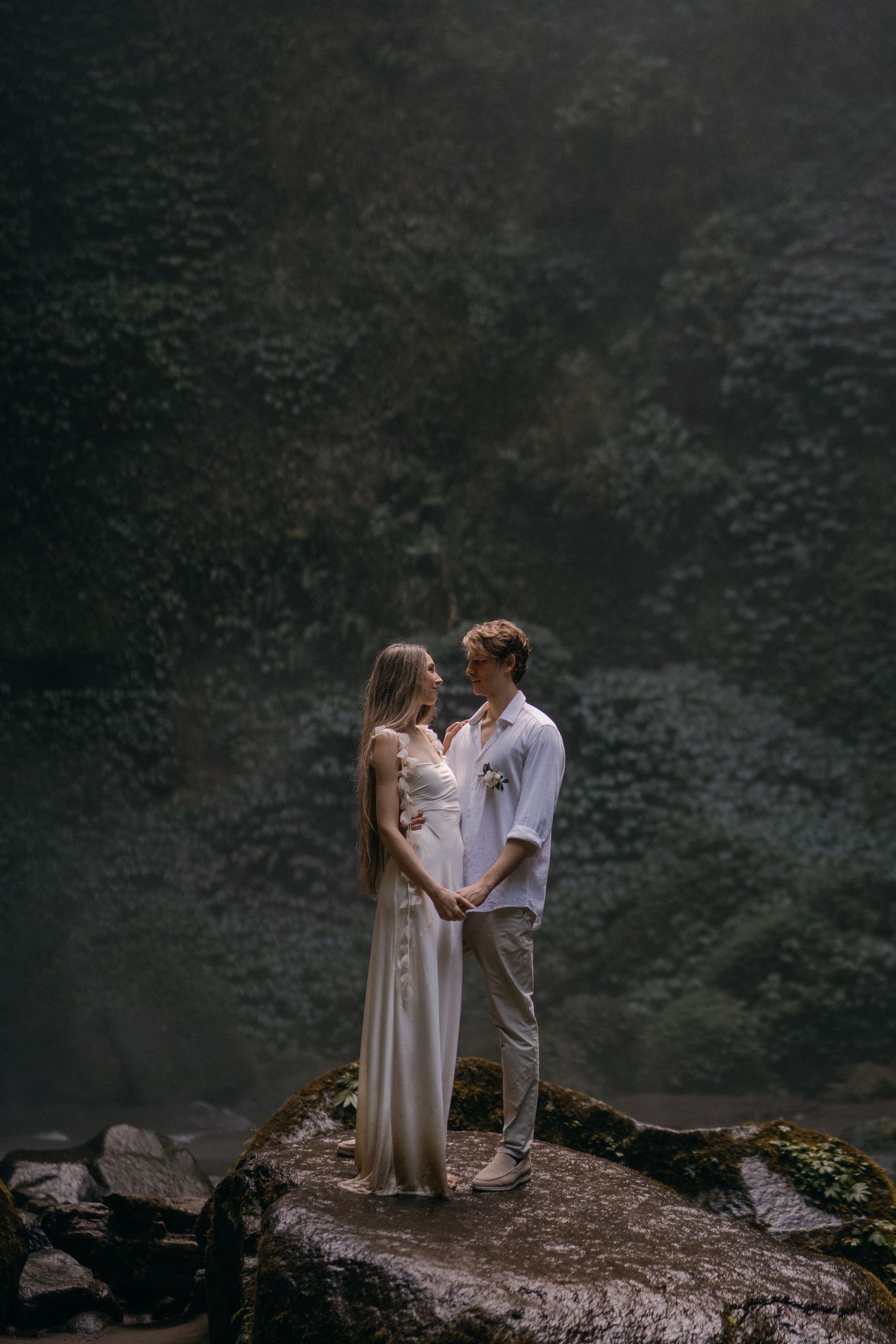 A couple in romantic embrace stands on a rock outcropping against a misty dark forest backdrop.