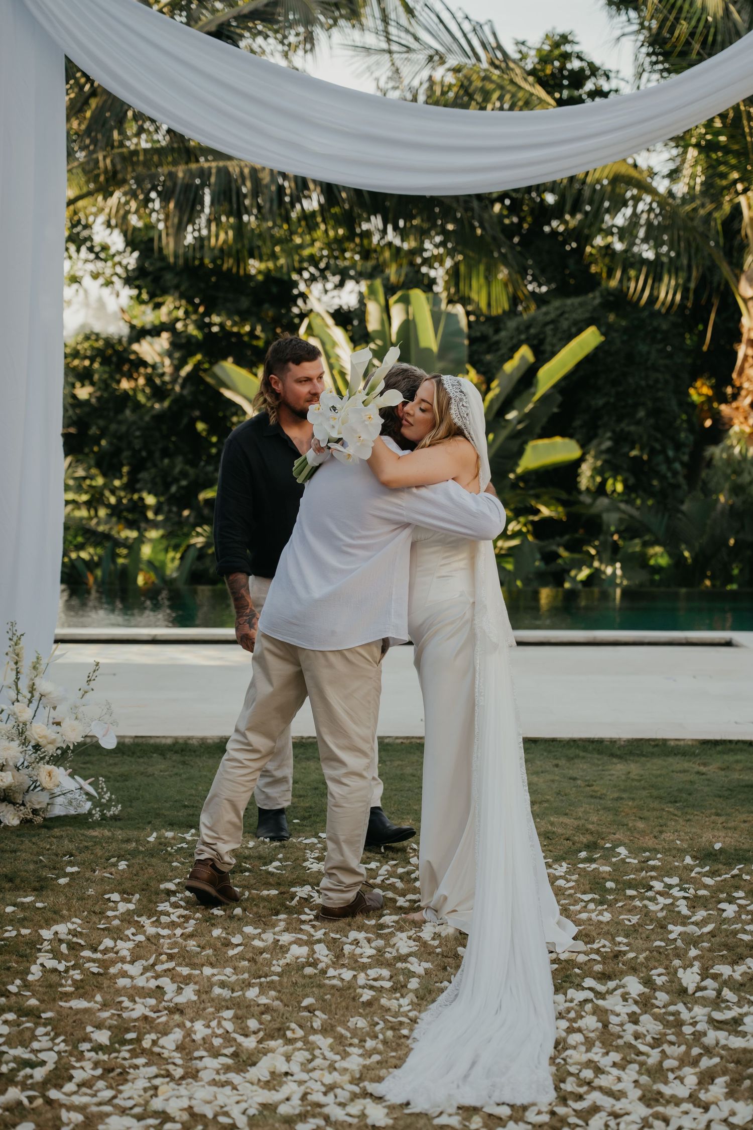 Bride in white gown embracing guest at outdoor tropical wedding ceremony with white arch.