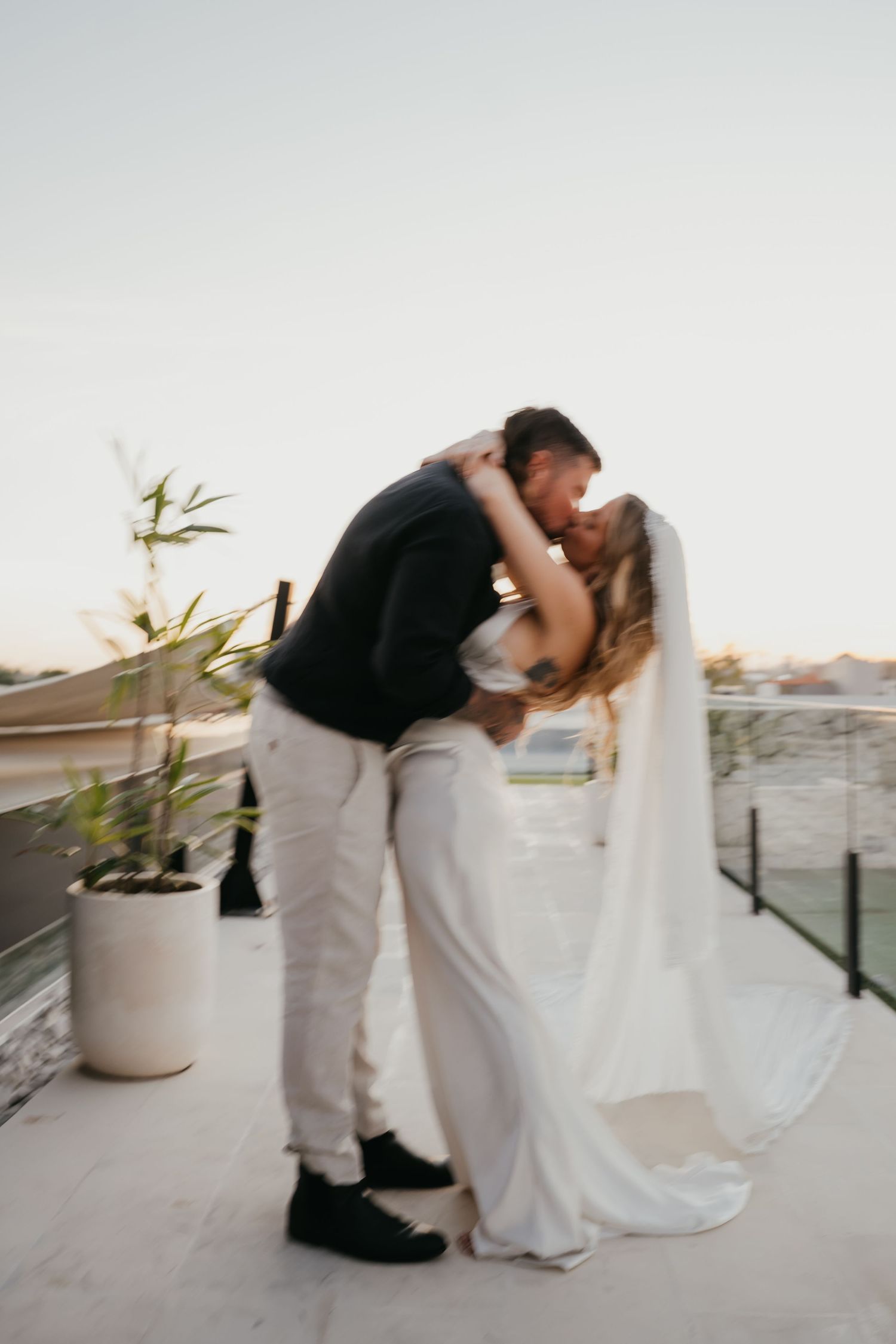 Groom dips bride for a kiss on rooftop terrace at sunset during romantic wedding portrait session.