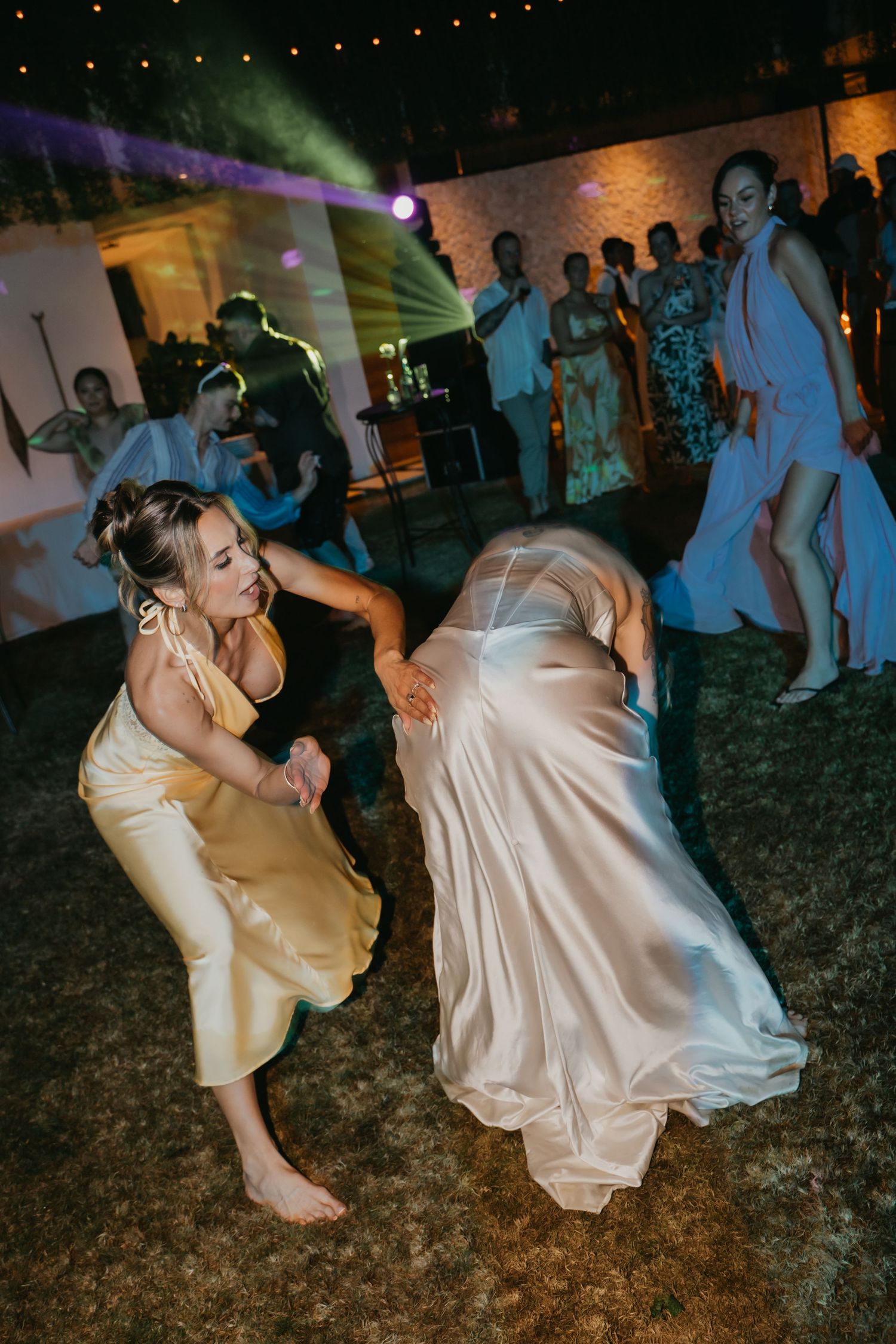 Bride in white satin gown dancing energetically with bridesmaid in yellow dress at reception.