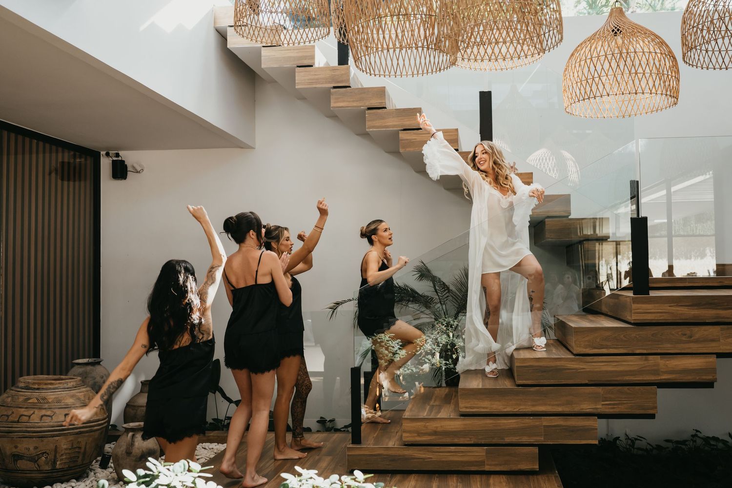 Women in black and white outfits celebrate on wooden stairs with rattan pendant lights during a bachelorette party.
