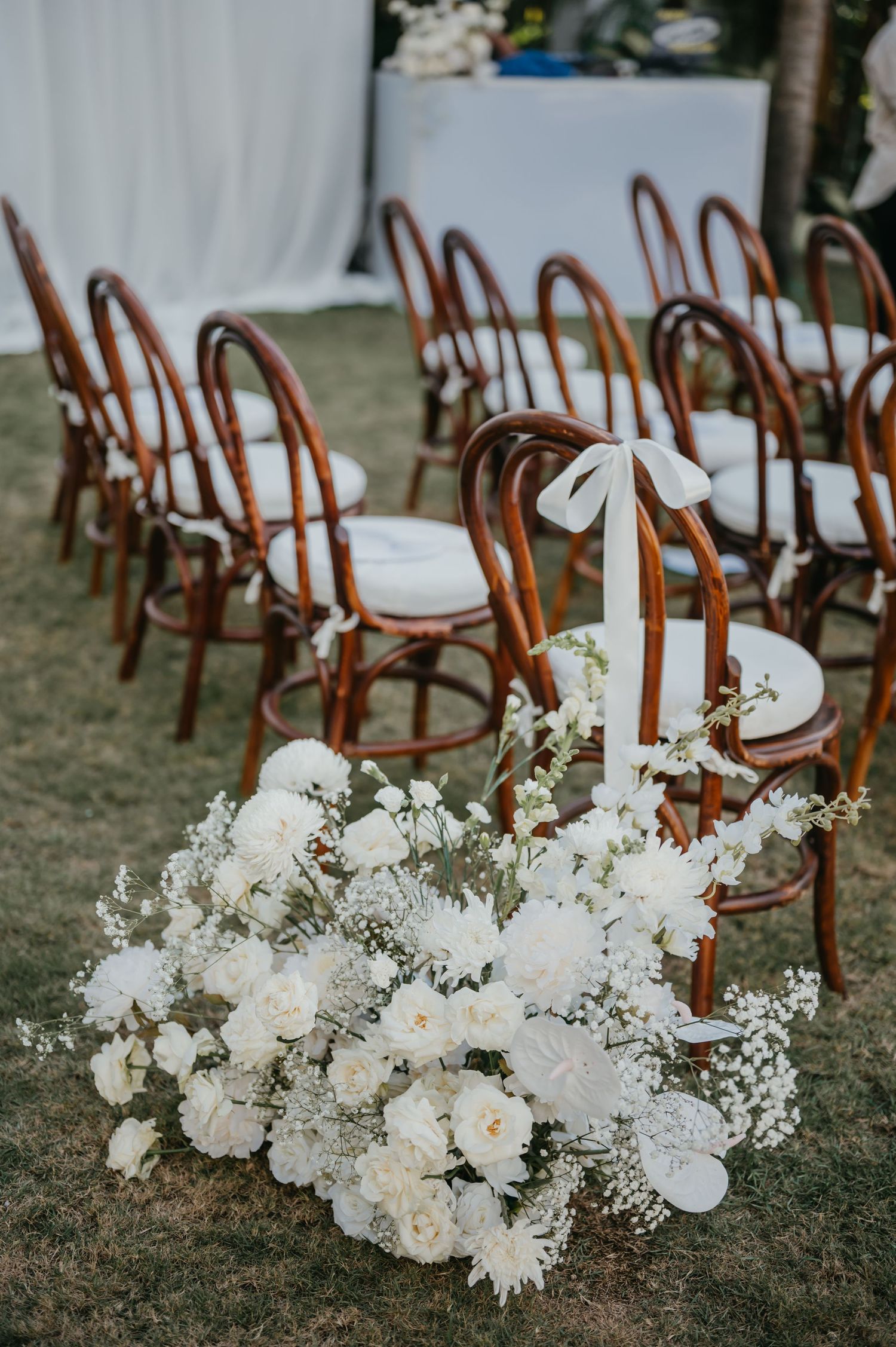 White floral arrangements line the aisle at an outdoor wedding ceremony with wooden chairs.
