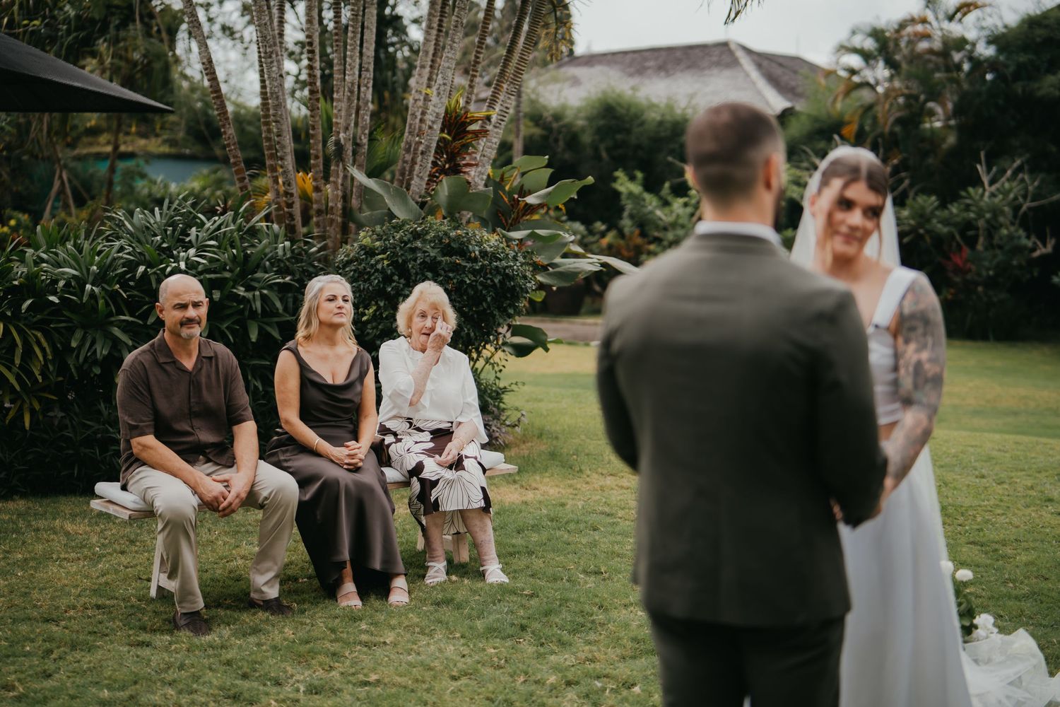 Groom and tattooed bride exchange vows before intimate group of seated guests in tropical garden setting.