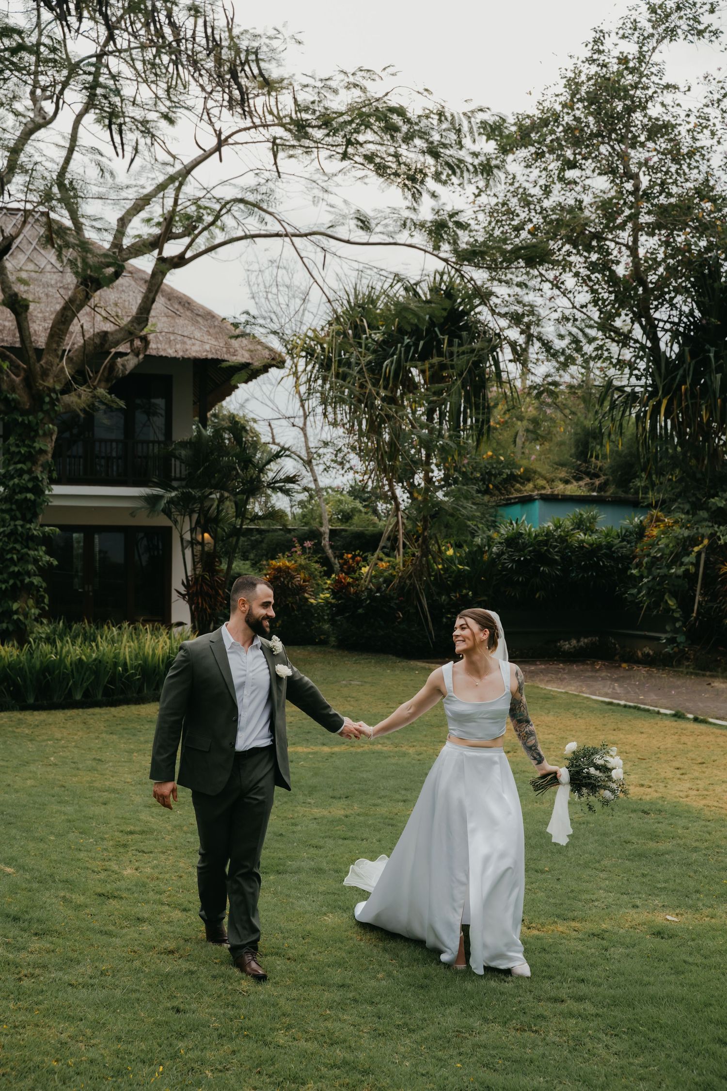 Bride and groom walk hand-in-hand across lush green lawn at tropical wedding venue surrounded by palm trees.