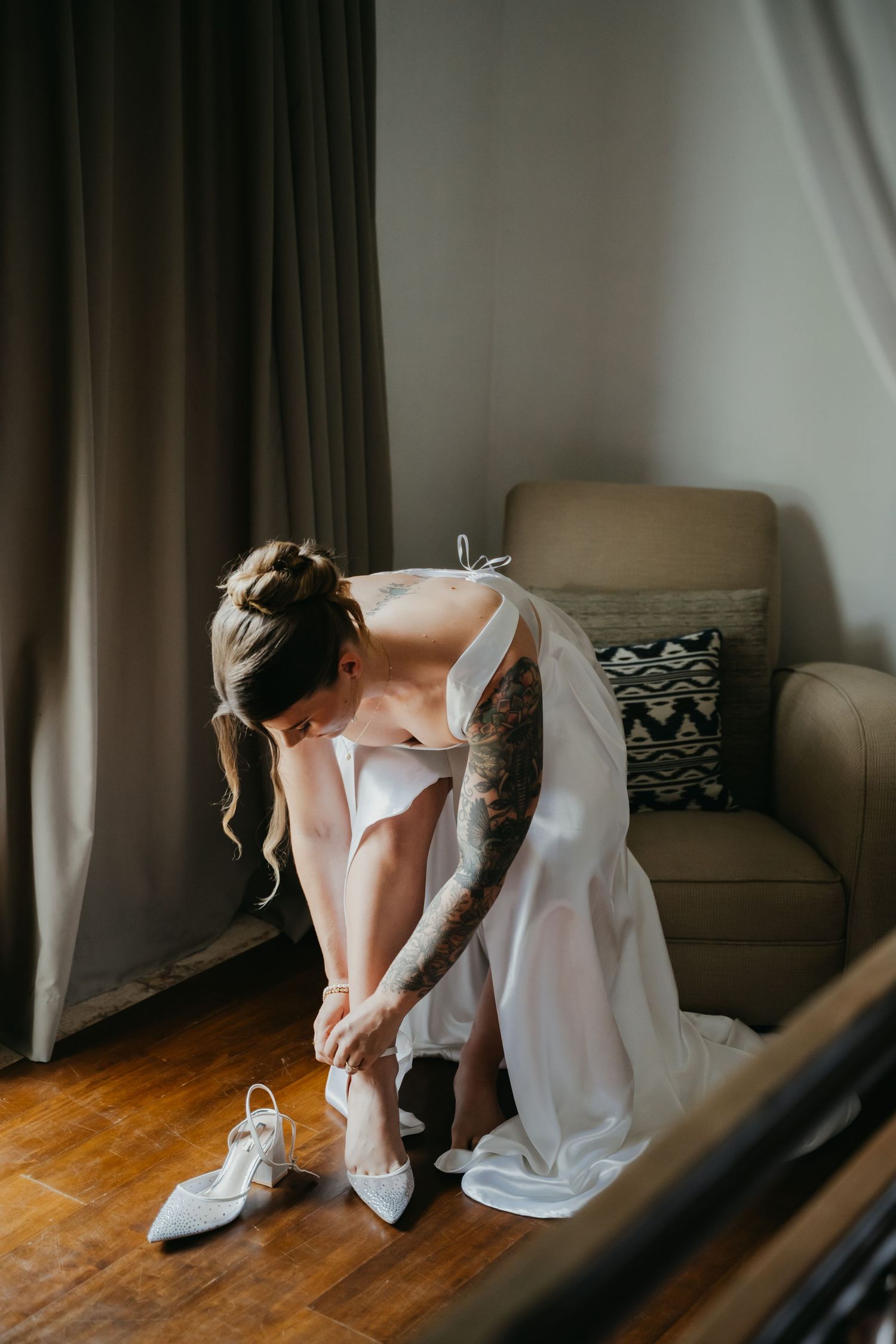 Tattooed bride in white wedding dress putting on heels while seated in armchair.