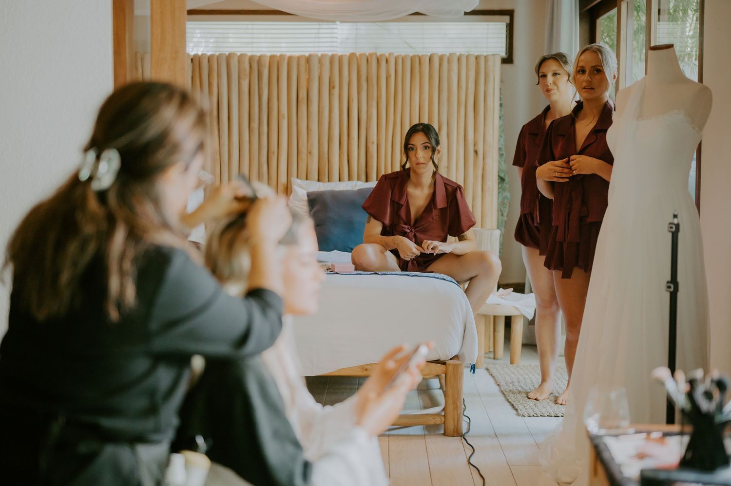 Bridesmaids in burgundy robes gather in a bright room near a wedding dress on a mannequin during bridal preparations.
