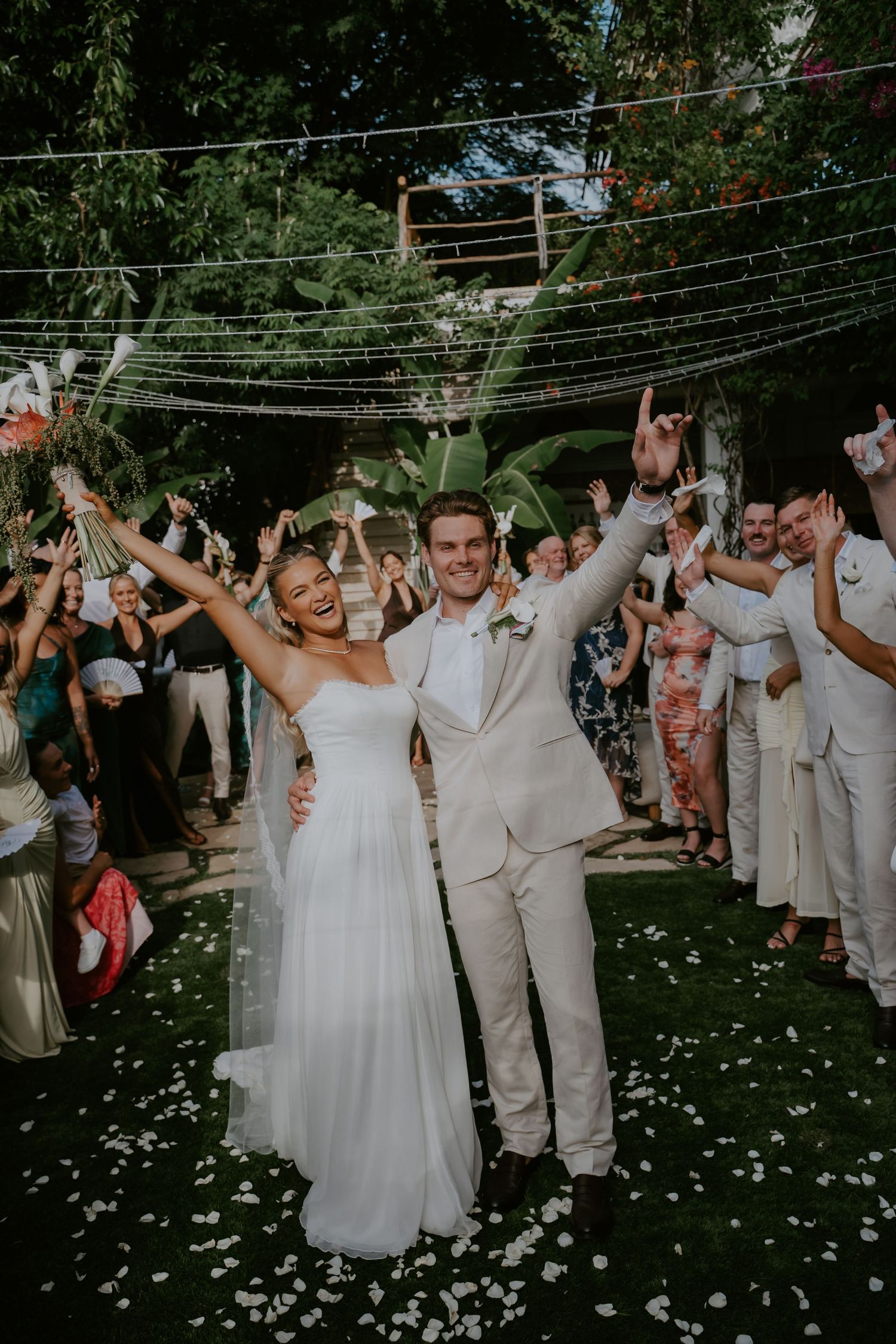 Joyful newlyweds celebrate recessional with raised arms as guests shower them with white flower petals.