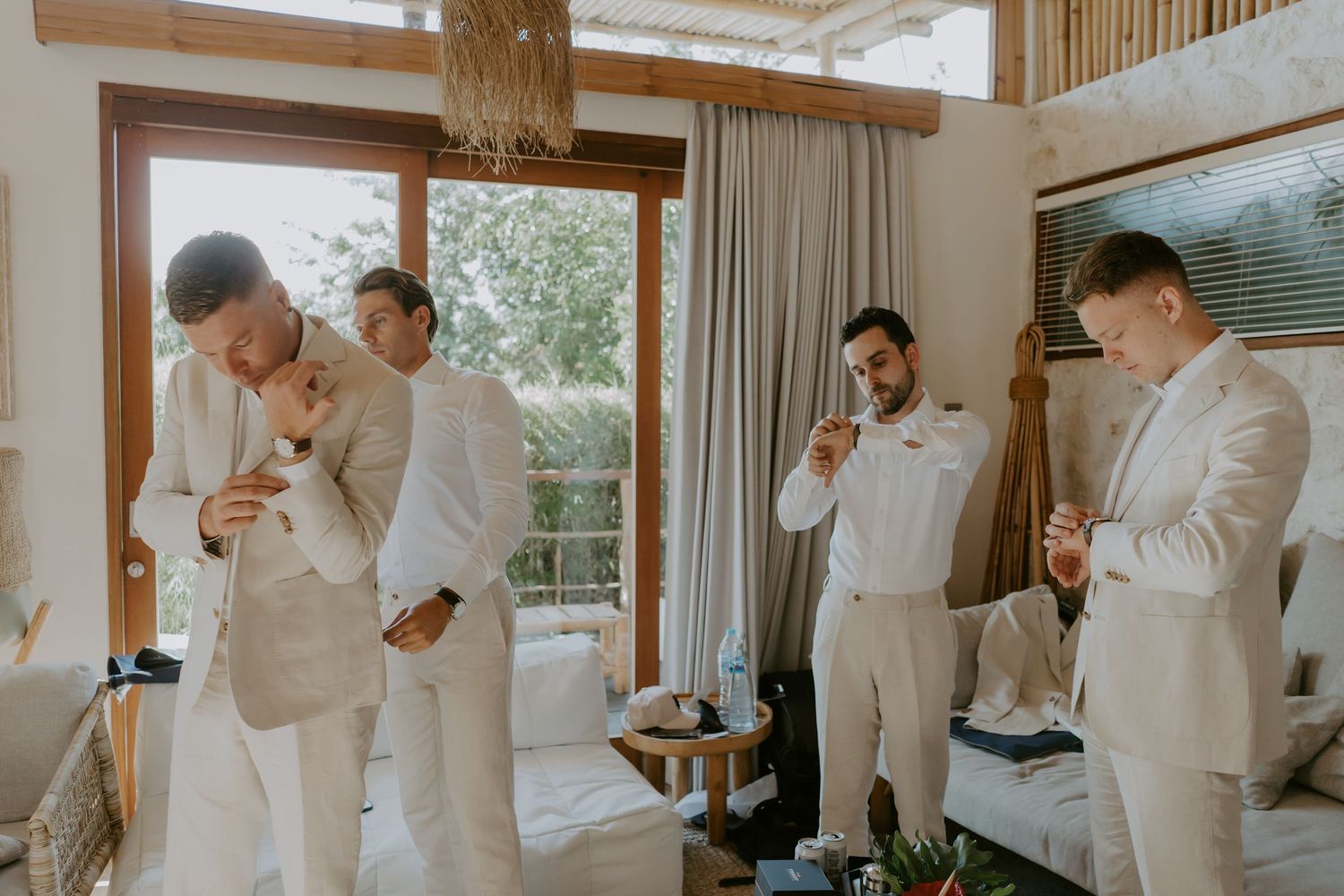 Groomsmen in cream suits helping each other get ready in a bright boho wedding suite.