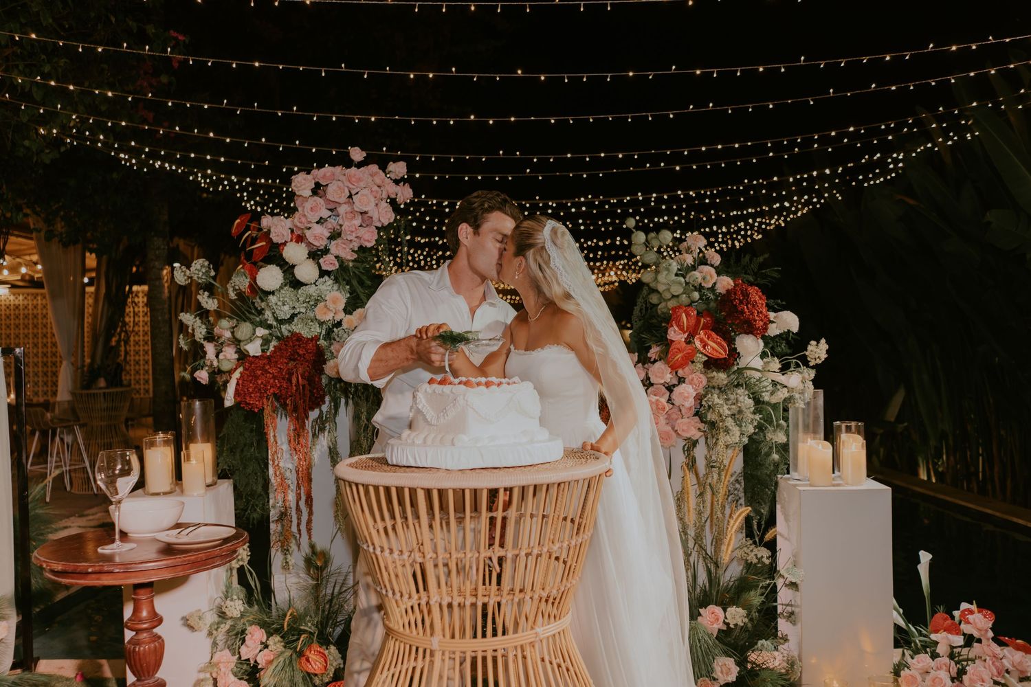 Newlyweds share a kiss over their wedding cake surrounded by candles and colorful floral arrangements.