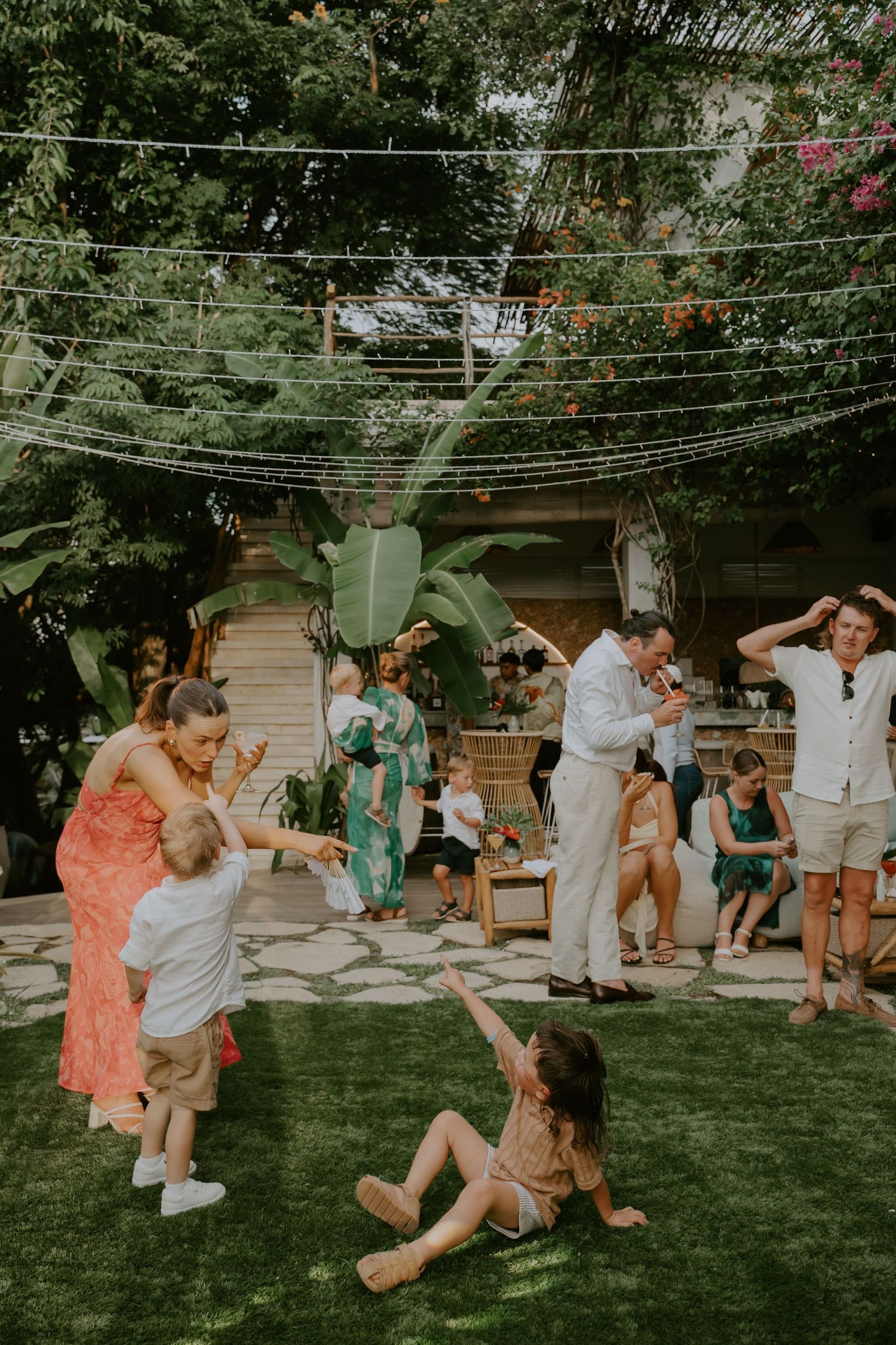 Wedding guests dance and play on green lawn during outdoor garden reception with string lights overhead.