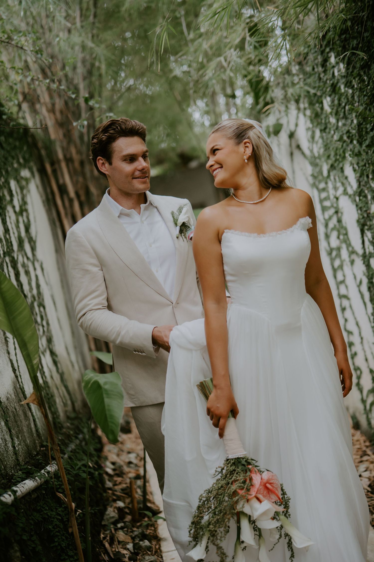 Smiling bride and groom walk hand-in-hand through a lush green garden pathway on their wedding day.