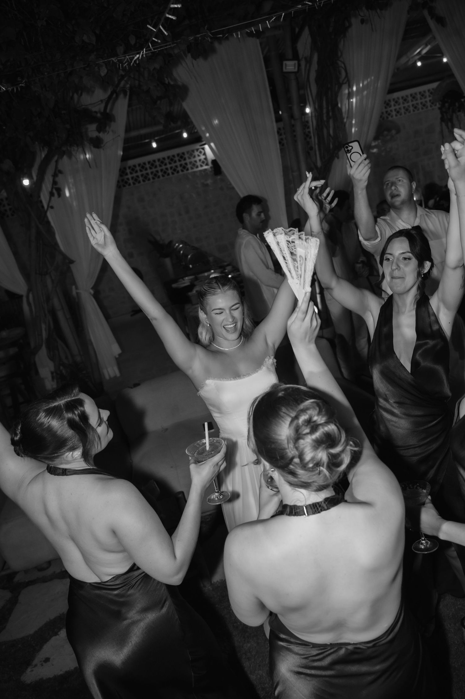 Black and white photo of bride dancing joyfully with bridesmaids at lively outdoor wedding reception.