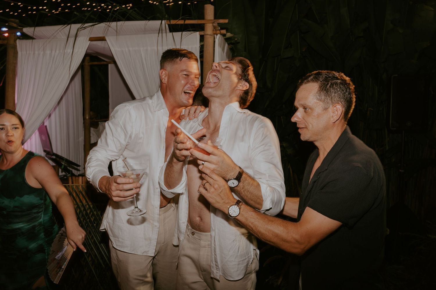 Three men laughing and toasting with cocktails during fun outdoor tropical wedding reception celebration.