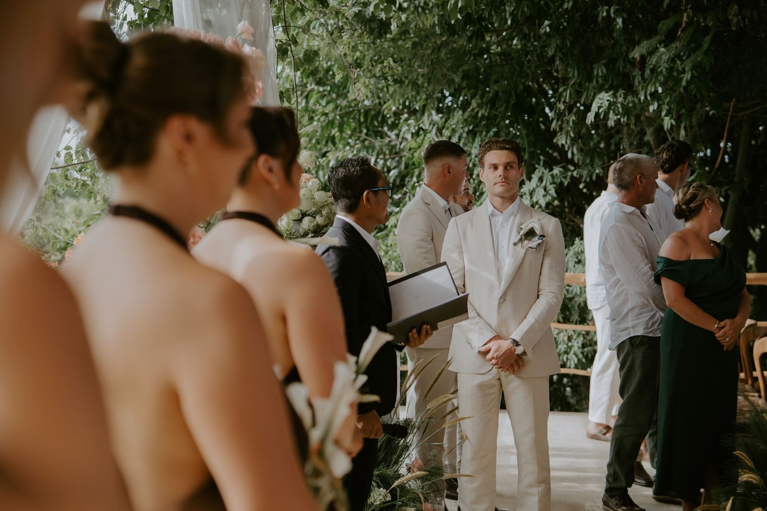 Groom in white suit watches bride approach at outdoor tropical wedding ceremony.