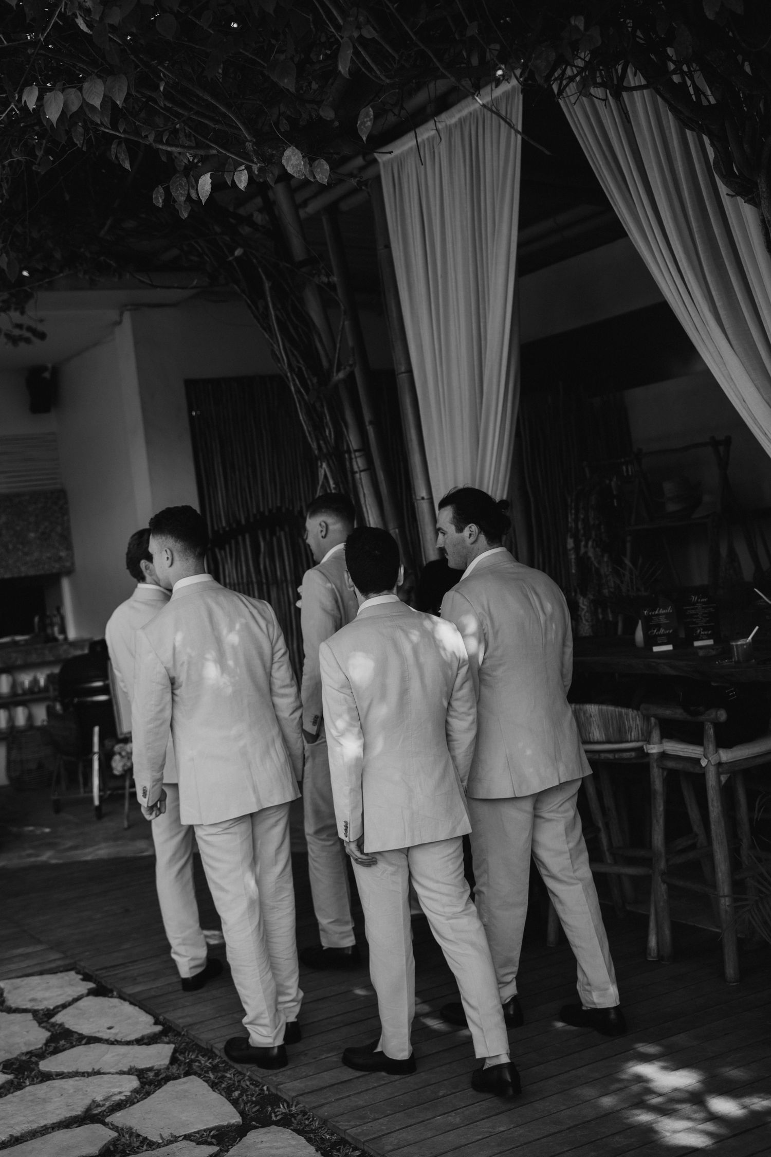 Groomsmen in white suits walk together toward a draped outdoor wedding venue.