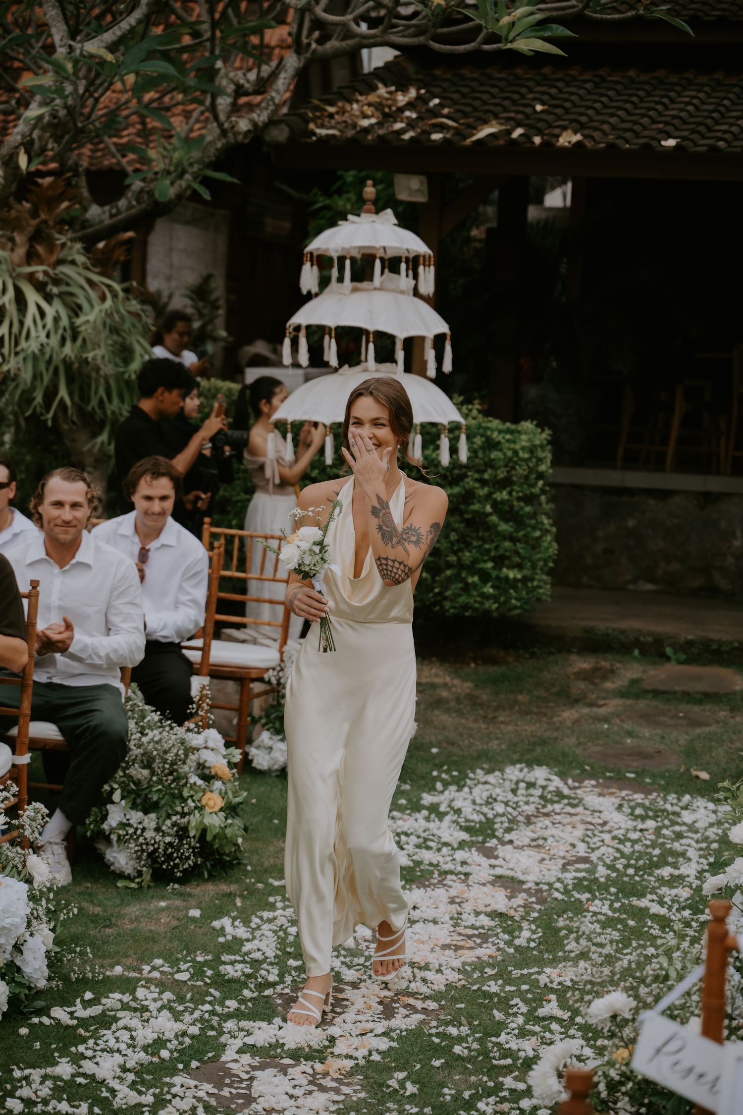 Emotional bride walks down petal-lined aisle holding umbrella and bouquet at outdoor garden wedding.